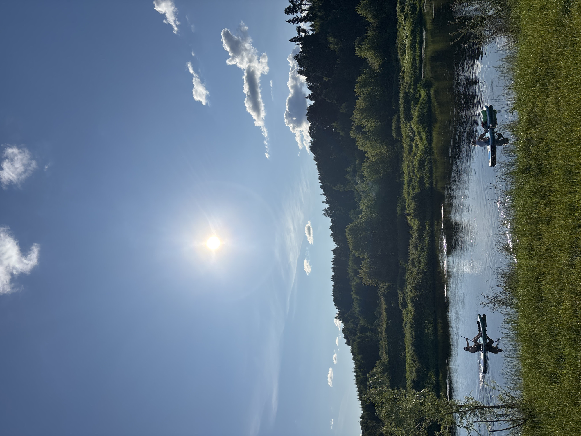 This is a vertical, medium-shot photograph taken on a sunny day. The scene depicts a wide, calm river reflecting the bright blue sky and a few scattered white clouds. Two individuals are visible on the water, each on a stand-up paddleboard. The person on the left is paddling, while the person on the right is seated. Both are silhouetted against the bright water. The riverbanks are lined with lush green grass and vegetation. In the background, a dense forest covers a gently sloping hill. The sun is high in the sky, casting a bright glare. The overall impression is one of a peaceful, natural environment. The image appears to have been taken in Zvenigorod, Russia, given the typical landscape.