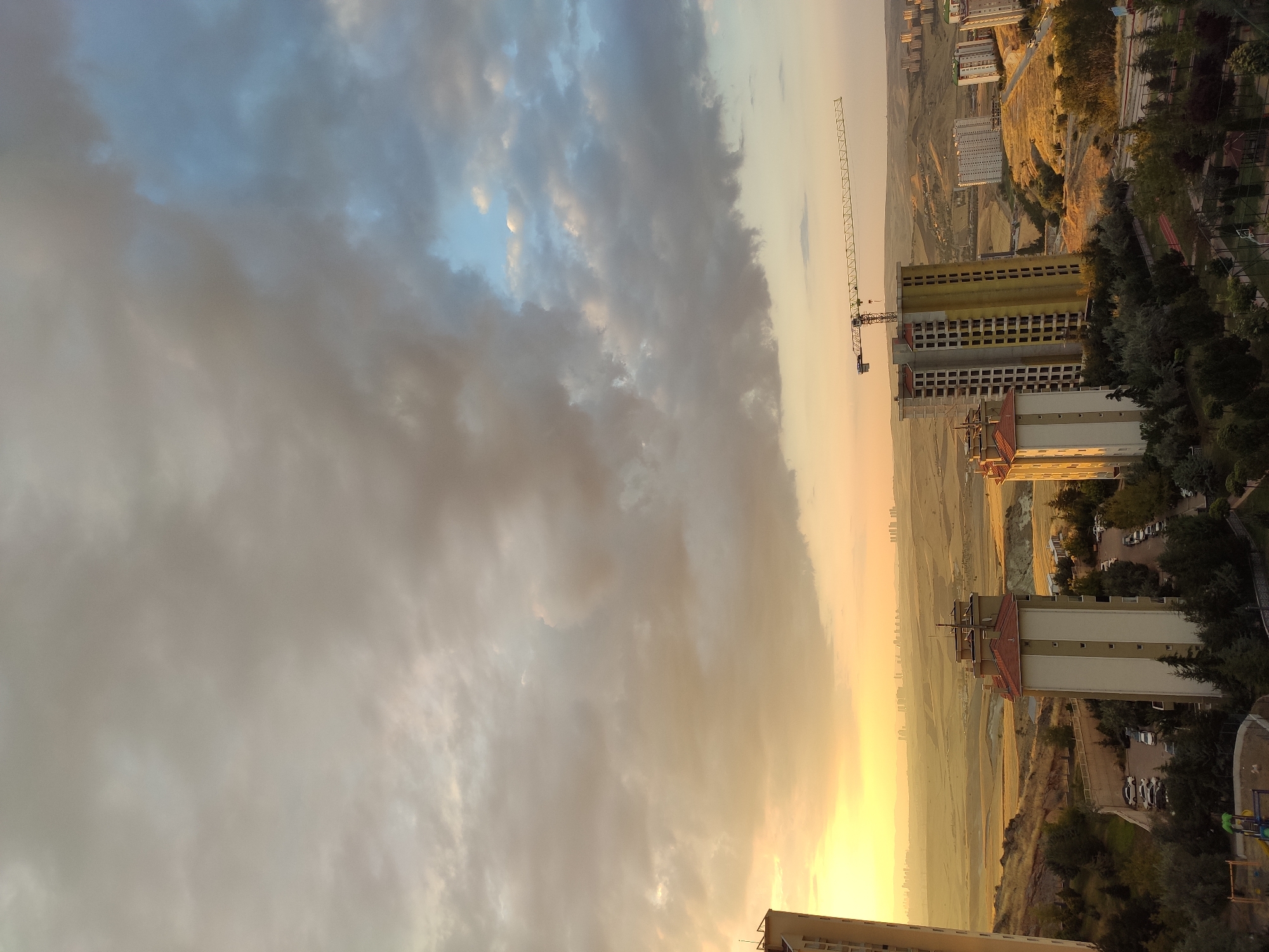 This image depicts an aerial view of the Etimesgut district of Ankara, Turkey, during what appears to be sunset or sunrise, given the warm, golden light illuminating the landscape and the dramatic, cloud-filled sky. In the foreground, several residential buildings are visible, some of which appear to be under construction, indicated by a prominent construction crane towering over one of the buildings. The buildings are varied in design, with some featuring red-tiled roofs and others having more modern, plain facades.
The middle ground is characterized by rolling, dry hills and fields, with some sparse vegetation and trees dotting the landscape. Further in the distance, a collection of urban buildings, likely forming a city skyline, is faintly visible through the atmospheric haze, suggesting the presence of a larger city beyond the immediate Etimesgut area.
The sky is a dominant feature, filled with a mix of heavy, dark clouds tinged with the warm hues of the setting or rising sun, and patches of clear blue sky. The overall impression is one of an urban development encroaching on a more natural, hilly terrain, under a dynamic and colorful sky. There are no visible people or animals in the frame. The scene is static, focusing on the architectural and natural elements of the Etimesgut district.
