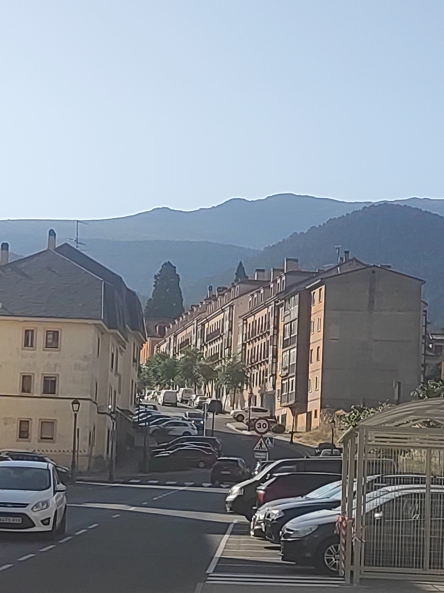 The image captures a street scene in the city of San Ildefonso, Spain, likely during daylight hours with clear skies. The perspective is from street level, looking uphill along a road lined with multi-story apartment buildings. The architecture suggests a European style, with balconies and many windows. On the left, a lighter-colored building with a dark mansard roof is prominent. On the right, a longer, narrower building with an orange-beige facade stretches down the street.
The street is lined with parked cars, indicating a residential or mixed-use area. Various makes and models of cars are visible, including a white Ford in the foreground on the left. Traffic signs are present, including a "30" speed limit sign and a pedestrian crossing sign. Trees and other greenery are interspersed between the buildings and along the roadside, adding to the urban landscape. In the background, rolling hills and mountains form a natural backdrop, their contours softened by the distance and atmospheric haze. The overall impression is one of a quiet, sunlit day in a European town or city. No people are clearly visible in the image, though the presence of parked cars suggests the area is inhabited.