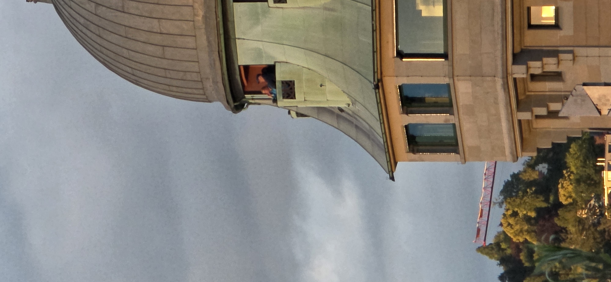 This image shows a close-up of the upper levels of a building with a domed roof, against a cloudy sky. The building is made of stone and has several windows. Two people are visible in a balcony area on the upper part of the building, looking out. The lower portion of the image shows trees and a red construction crane in the distance. The lighting suggests it is either early morning or late afternoon.