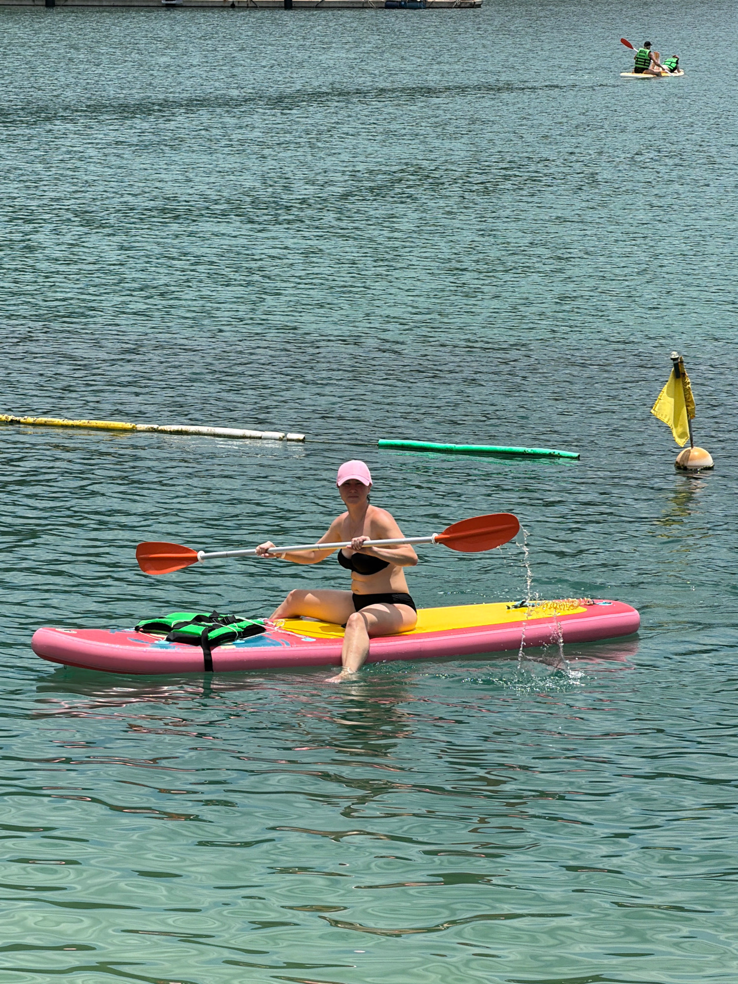 A woman in a pink cap and black bikini sits on a paddleboard in clear, turquoise water. She holds an orange-bladed paddle, mid-stroke, creating a splash. The paddleboard is pink and yellow with a green life vest strapped to the front. In the background, another paddleboard with two people wearing life vests is visible. A yellow flag on a buoy and a long green float are also present in the water. The scene appears to be a waterfront area, likely a beach or bay in Nha Trang, Vietnam, given the clarity of the water and the presence of recreational water activities.