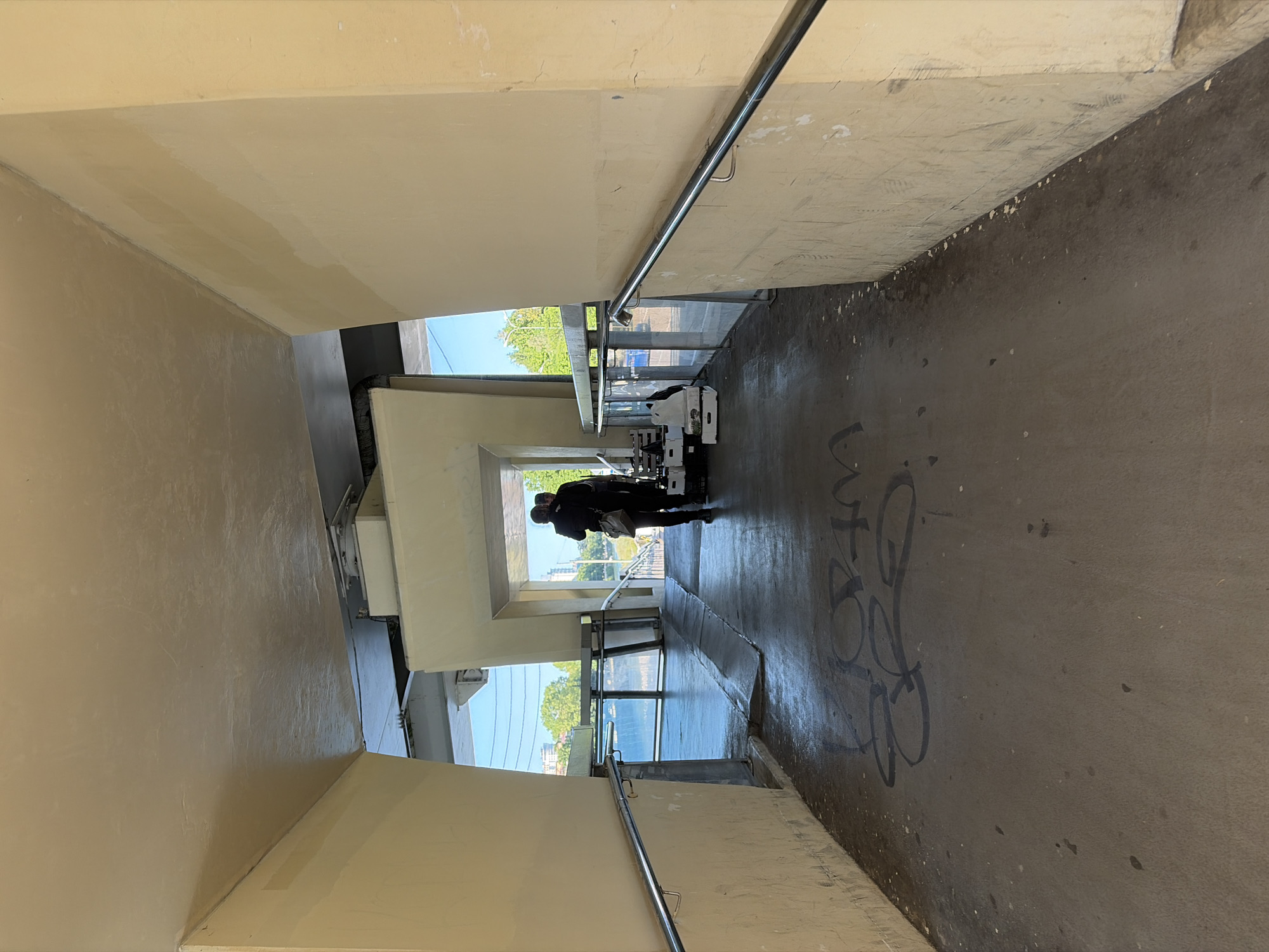 The image displays a concrete pedestrian underpass or walkway, viewed from a slightly elevated position looking downwards along its length. The photograph is oriented vertically, rotated 90 degrees counter-clockwise. The underpass features light-colored, possibly painted, concrete walls and ceiling, contrasted with a darker, grimy concrete floor. Metal handrails are present along both sides of the pathway, suggesting a ramp or set of stairs.
Two individuals are visible in the distance, walking away from the viewer towards the exit of the underpass. They are silhouetted against the bright light from outside. One person appears to be pushing a trolley or cart, while the other walks alongside. Both are dressed in dark clothing.
The exterior beyond the underpass reveals a clear blue sky and green trees, indicating it is daytime with sunny weather. Light filters into the underpass from these openings, illuminating the distant sections more brightly than the foreground. The floor in the foreground shows signs of wear, with scattered dark spots and a prominent piece of black graffiti featuring abstract, looping letters, although no specific words are clearly legible. The walls also exhibit some marks and discoloration. The scene depicts a functional, somewhat worn public thoroughfare in Sochi, Russia.