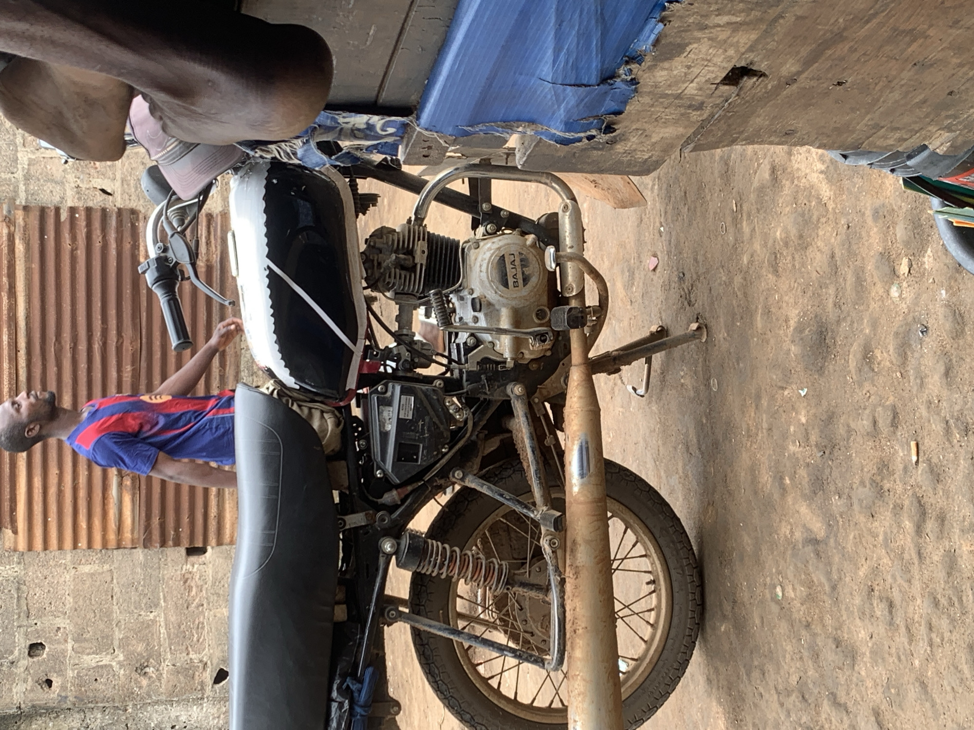 In the city of Shagamu, Nigeria, a man in a blue and red striped t-shirt walks past a black and white motorcycle. The motorcycle's engine is visible, bearing the "BAJAJ" logo. The rear wheel of the motorcycle is muddy, as is the exhaust pipe and surrounding ground. A corrugated metal fence is visible in the background, behind a rough brick wall. To the right of the motorcycle, a section of a blue tarp covers a wooden structure, with a dark-colored bucket visible below.