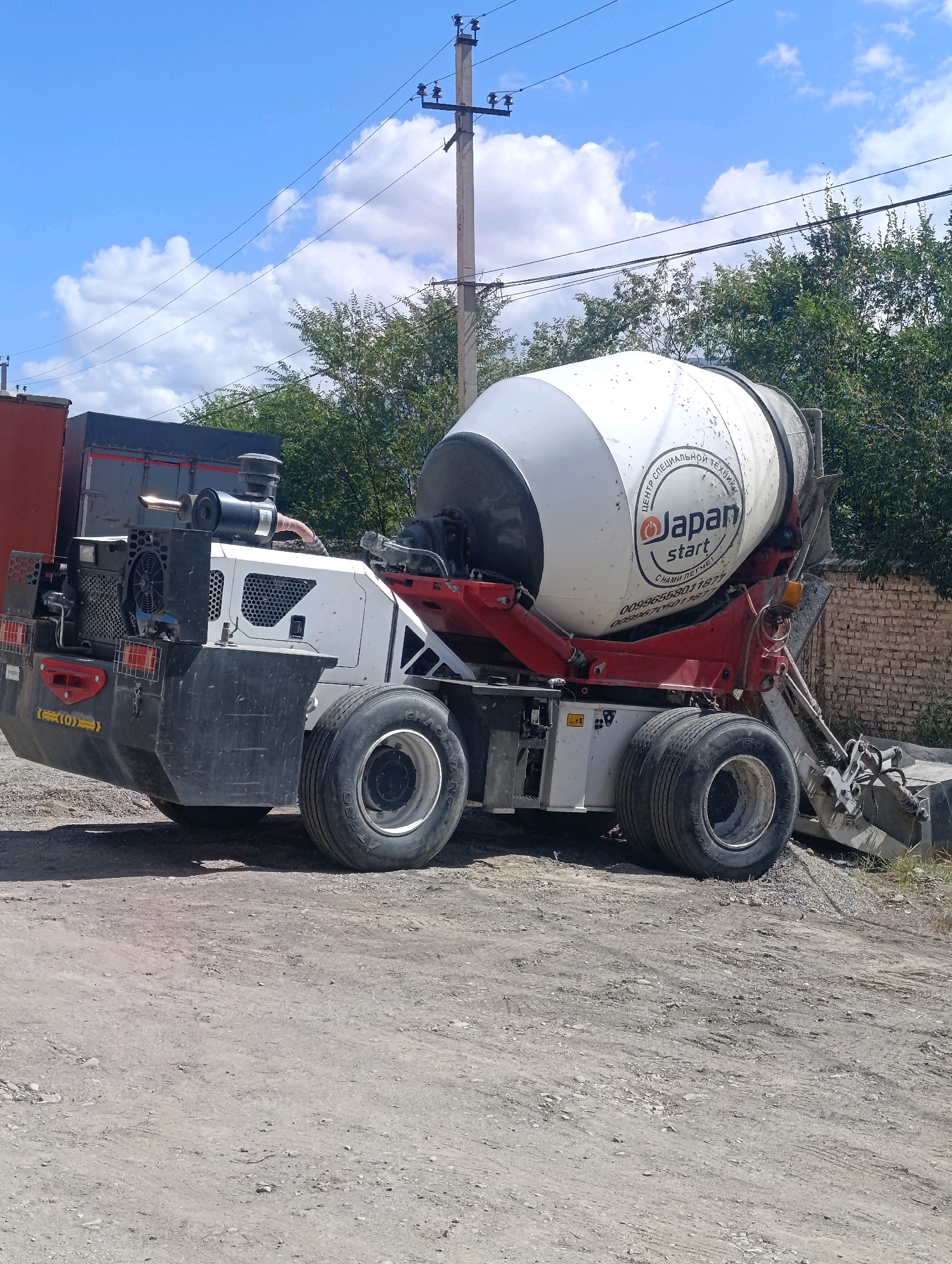In Naryn City, Kyrgyzstan, a concrete mixer truck with a white and red drum is parked on a dirt surface next to a brick wall and green trees. The truck has a black rear section with red taillights and large tires. The drum of the mixer has a circular logo that reads "Japan start" in English, along with Russian text above and below it. The sky is blue with scattered clouds.