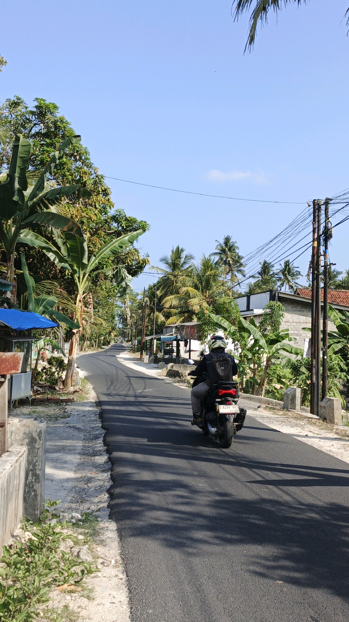 A person rides a motorcycle on an asphalt road in a rural area, likely in Cintaratu, Indonesia. The rider, wearing a helmet and a backpack, is moving away from the camera. The road is flanked by vegetation, including banana trees and palm trees, and a few buildings are visible in the background. Power lines crisscross the sky. The lighting suggests it is daytime, with shadows cast on the road by the trees.