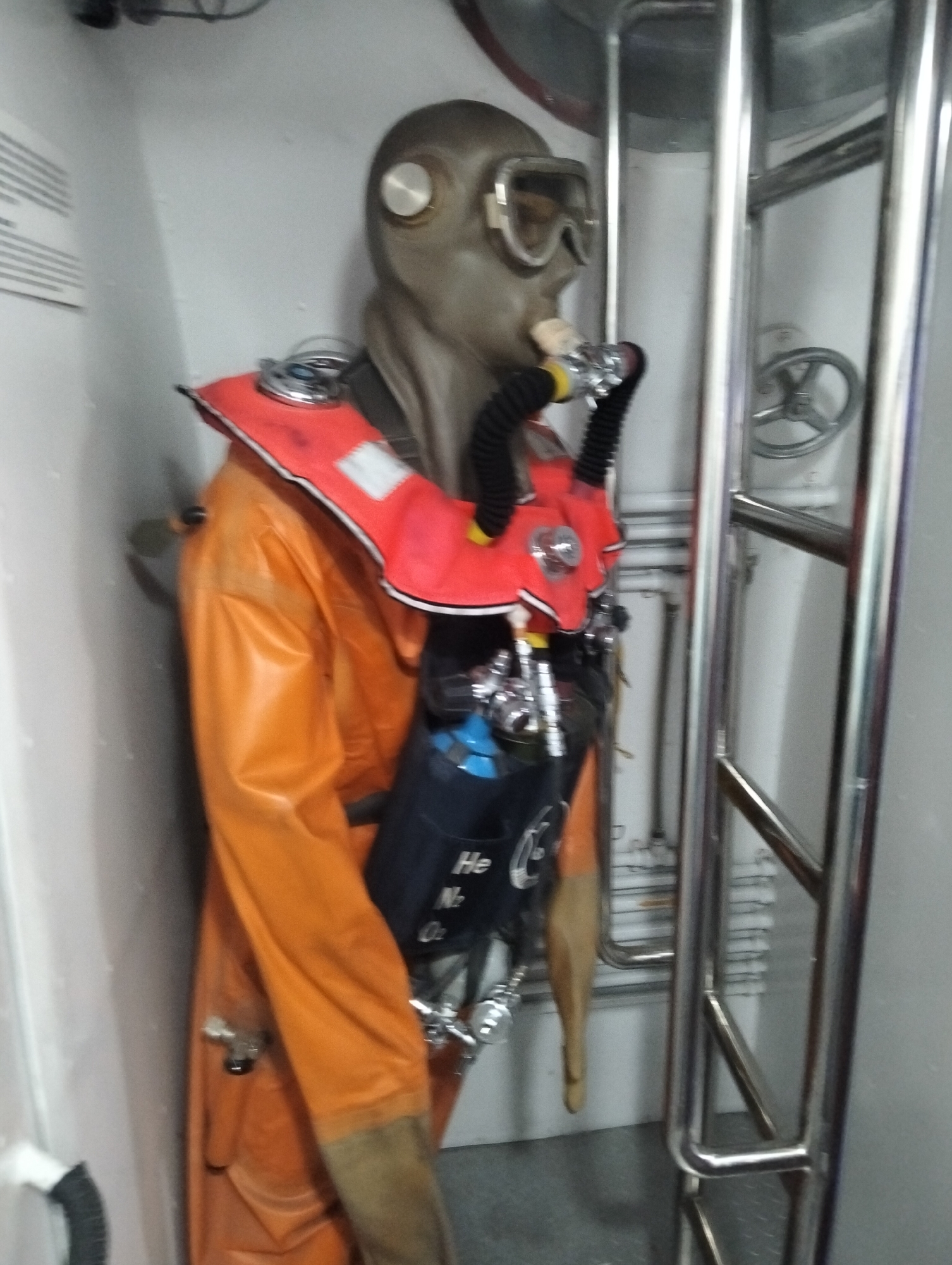 The image shows a mannequin dressed in a deep-sea diving suit. The suit is orange, and the diver has a grey full-face mask with goggles, and a red flotation device around the neck. A black and blue diving tank is strapped to the mannequin's back. The mannequin is standing next to a metal ladder in what appears to be an enclosed space, possibly the interior of a ship or submarine. There is a metal valve visible on the wall behind the ladder. The specific location in Dhaka, Bangladesh is not discernible from the image.