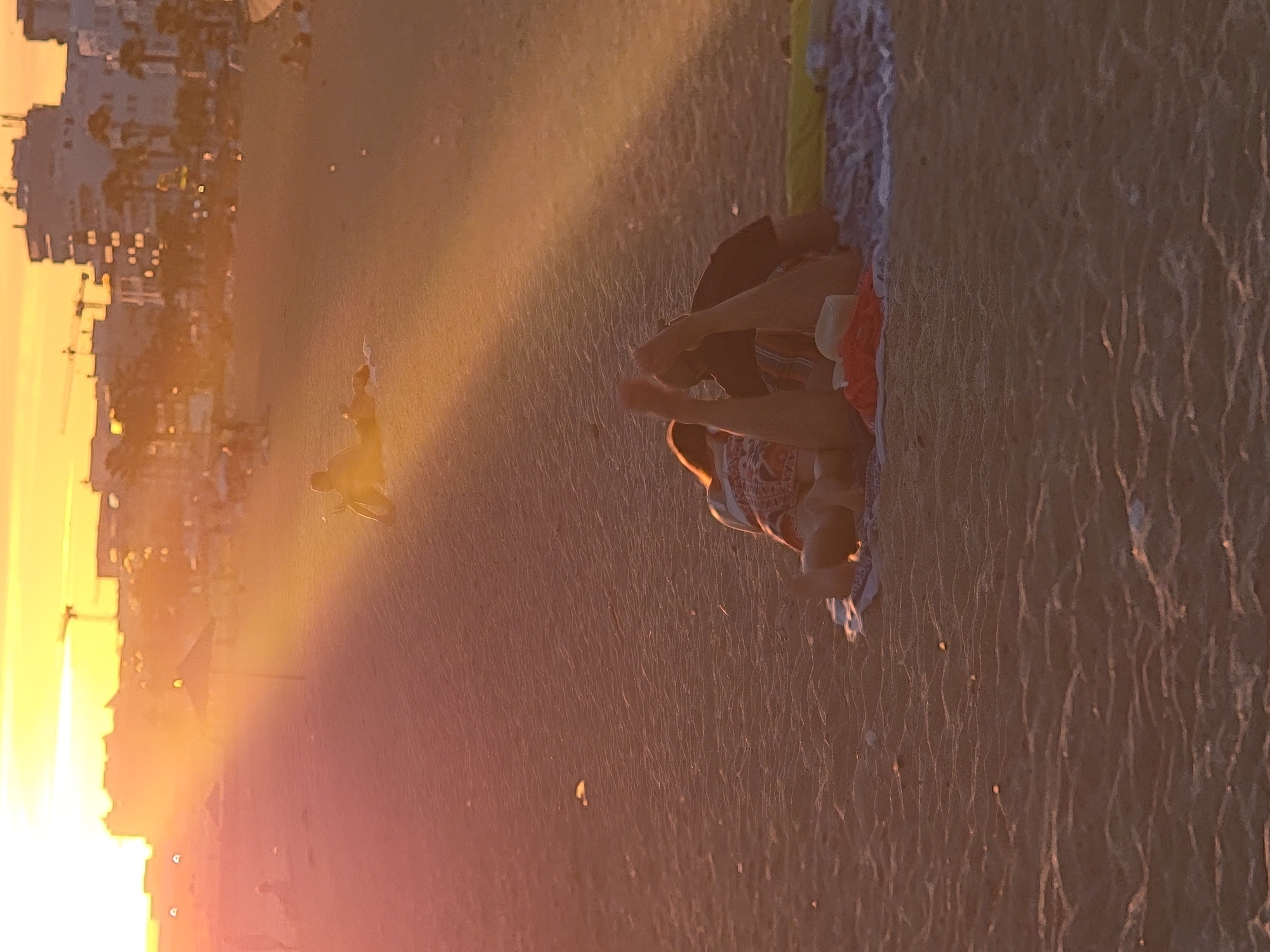 The image depicts a sunset scene on the beach in Quarteira, Portugal. In the foreground, two individuals are lying on blankets on the sand. One person, appearing to be wearing a patterned top and shorts, is partially obscured. The other person, wearing a dark shirt, is lying beside them. The legs of both individuals are visible, with one person's feet raised. A straw hat and a red garment are placed on the blanket. In the mid-ground, other beachgoers are scattered across the sand, some sitting and others lying down. Further in the background, buildings and palm trees are visible against the bright, hazy sky, with construction cranes also present, indicating urban development. The sunlight casts long shadows across the sand, creating a warm, golden glow over the entire scene.