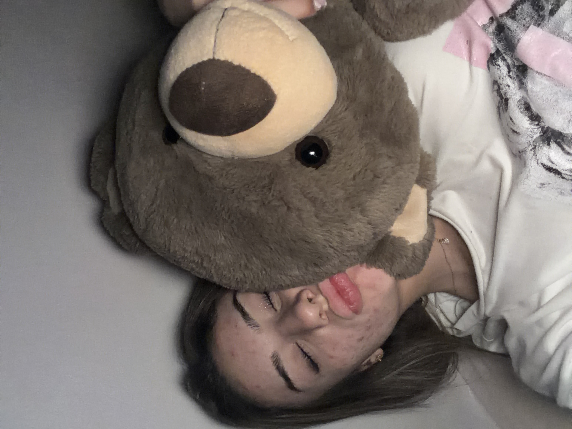 A young woman with closed eyes and her lips puckered is embracing a large stuffed teddy bear. She is wearing a white t-shirt with a graphic print. The background is a plain, light-colored wall. The scene appears to be indoors. There is no indication of the city of Shumen, Bulgaria in the image.