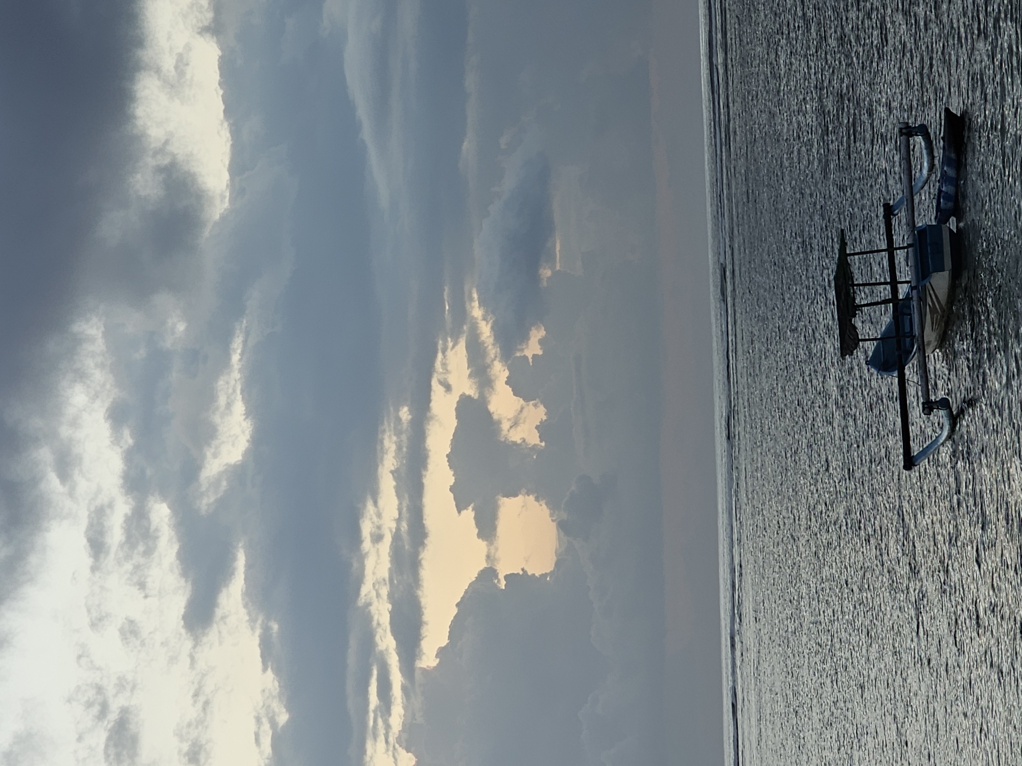 An overcast sky with scattered clouds in varying shades of gray and white hangs over a calm ocean. A narrow opening in the clouds reveals a pale orange glow, suggesting either sunrise or sunset. In the foreground, a traditional Indonesian outrigger boat, known as a jukung, is anchored on the water. The jukung is painted white and blue with a dark canopy supported by wooden poles. A smaller, dark blue and white striped boat is tethered to it. The ocean surface is rippled and reflects the muted light of the sky. The horizon line is clear, with faint waves breaking in the distance. The scene appears to be a tranquil moment at sea, possibly near the coast of Denpasar, Indonesia, given the presence of a jukung.