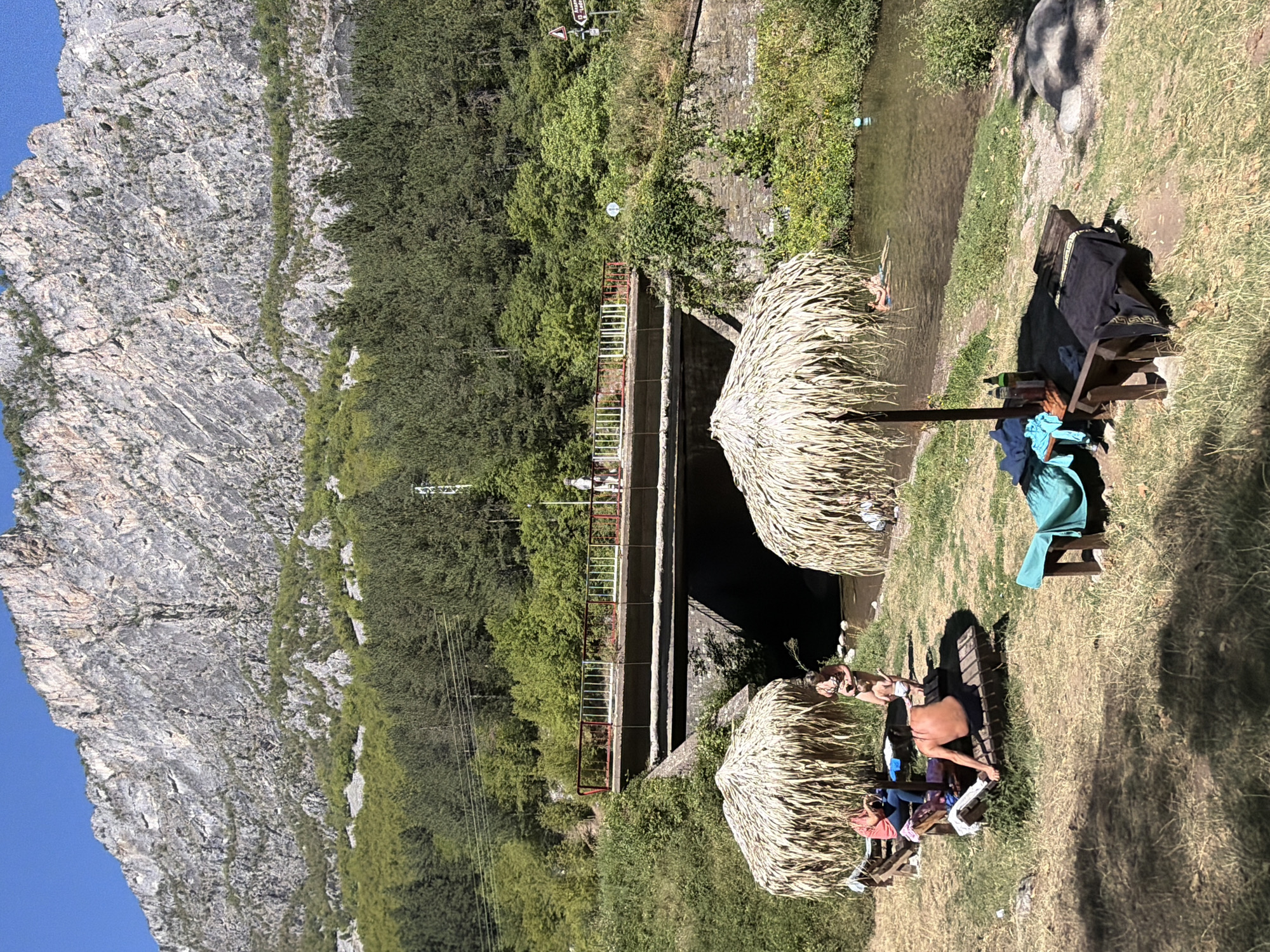 The image depicts a scenic outdoor area near Zgorigrad, Bulgaria, characterized by a river, a bridge, and a mountainous backdrop. In the foreground, a grassy bank borders a clear river where a few people are visible swimming. Several wooden lounge chairs and two thatched umbrellas are set up on the grass, suggesting a recreational spot. On one lounge chair, a person is reclining. Nearby, a group of three individuals are gathered, with one person sitting and two standing. A bridge spans the river in the midground, with a few people walking on it. The background features a dense forest that climbs up the side of a rugged, rocky mountain under a clear blue sky. The overall scene conveys a tranquil summer day in a natural setting.