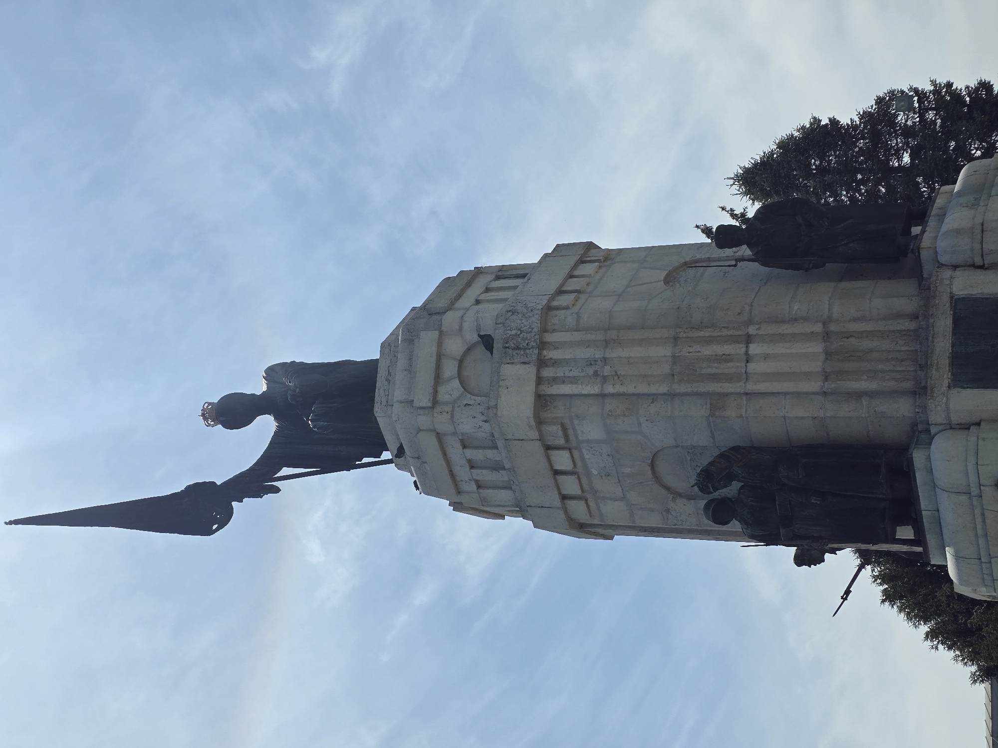 The image depicts a monument in an unidentified city. The monument features a large, multi-tiered, light-colored stone base, possibly made of marble or a similar material. Bronze statues are positioned on the base, appearing to depict figures with possible weapons. At the top of the monument stands a large bronze statue of a figure, likely a female figure, holding a banner or flag. The statue is silhouetted against a cloudy sky. Trees are visible adjacent to the monument, suggesting a park or public space setting. The angle of the photograph is from a low perspective looking upwards at the monument.