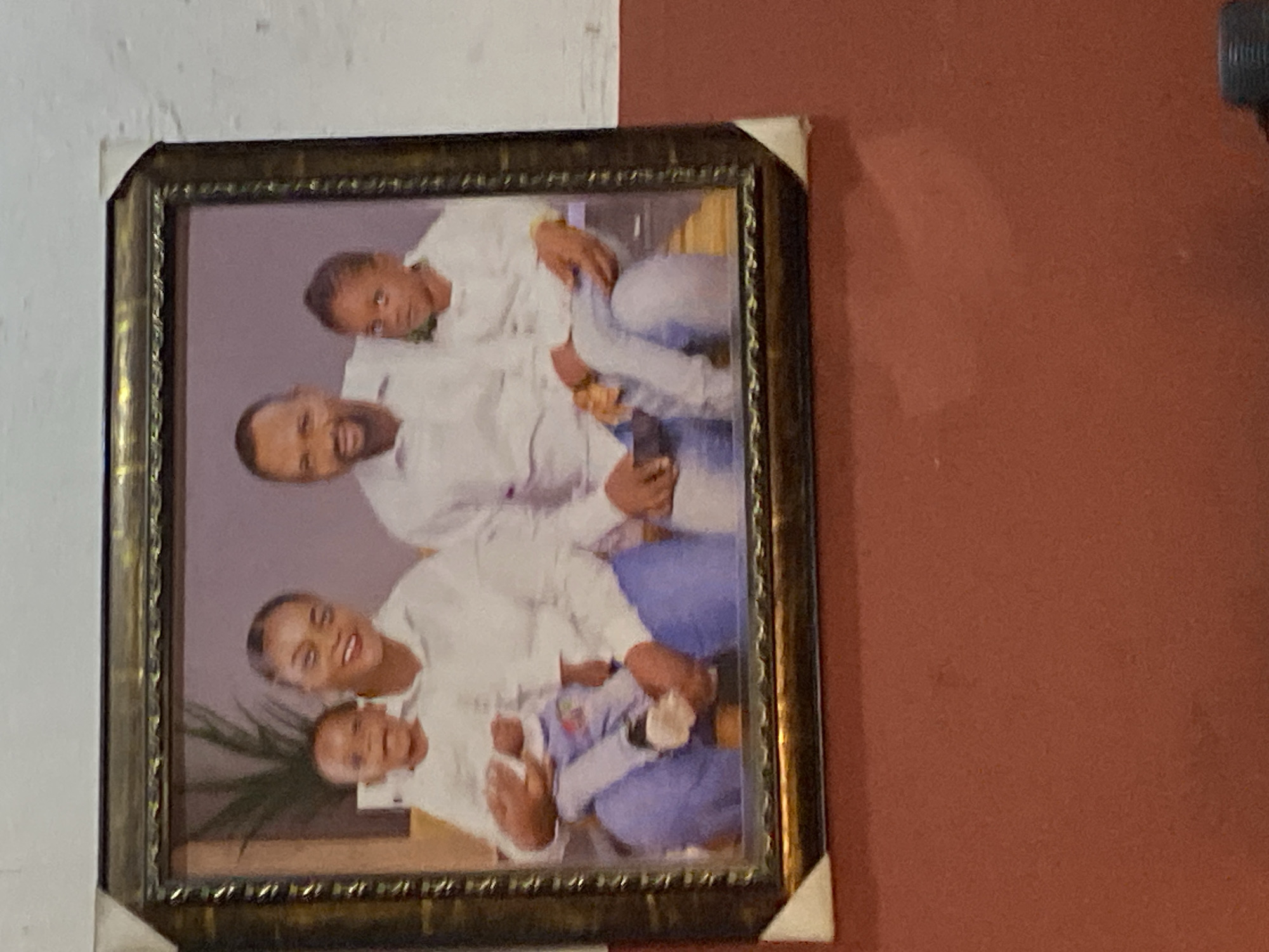 A medium close-up image displays a framed family portrait hanging on a wall, rotated 90 degrees clockwise. The ornate frame is dark gold or bronze with intricate detailing, and two white corner protectors are present on its top-left and bottom-right corners. The wall behind the frame has two distinct sections: the upper left is textured white or off-white, while the larger section to the right is a solid reddish-brown or terracotta.
The family portrait inside the frame features five individuals of Black African descent: two adults (one male, one female), two older children (appearing to be a boy and a girl), and an infant. All members are dressed in matching white tops and light blue bottoms, likely jeans. They are posed closely together, smiling warmly at the camera, conveying a sense of happiness and unity. The infant is held by one of the adults. The background of the portrait itself is a soft purple or lavender hue, with the partial view of a green leafy plant visible in the lower-left corner. The setting is indoors, characteristic of a home environment, possibly in Mararaba, Nigeria. No text is visible in the image.