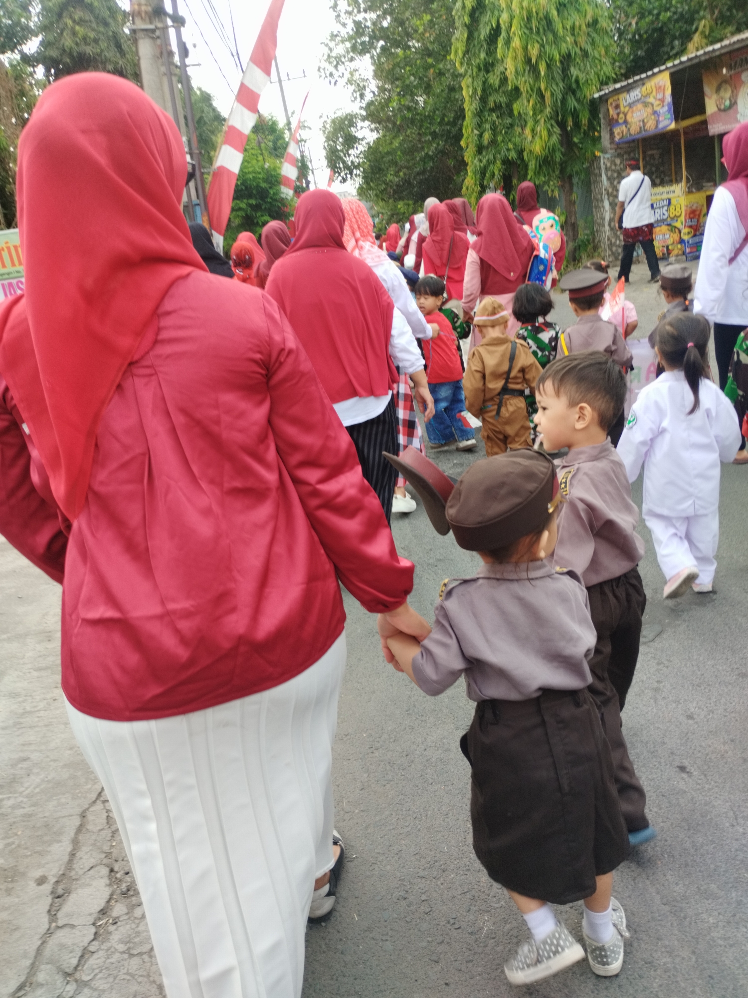 In Surabaya, Indonesia, a group of children and adults are walking on a paved street. The children are dressed in various costumes, including what appear to be police uniforms and traditional Indonesian attire. Some of the children are wearing brown hats and shirts with badges, while others are in white uniforms. There are also adults present, some wearing red headscarves and matching red tops. Red and white Indonesian flags are visible in the background, suggesting a festive occasion or national celebration. The environment appears to be outdoors, with trees and a building with signage visible. The overall scene depicts a cheerful procession or event involving children.