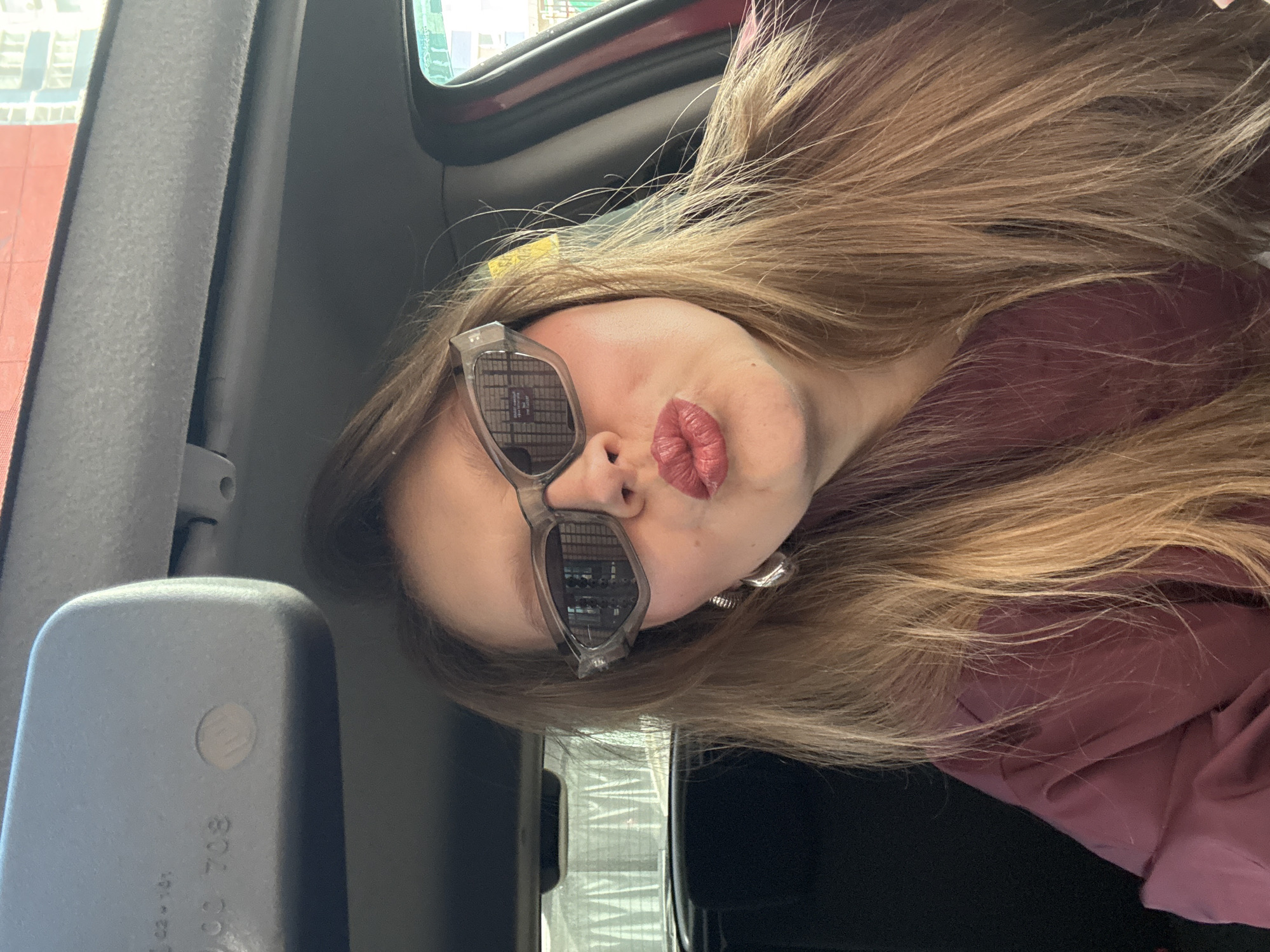 A woman with long, light brown hair is posing for a selfie inside a vehicle. She is wearing sunglasses with a building reflected in the lenses and has her lips puckered. The interior of the vehicle is visible, including a side mirror, a gray pillar, and part of the window frame. Reflected in the sunglasses lenses is a building in Izhevsk, Russia.
