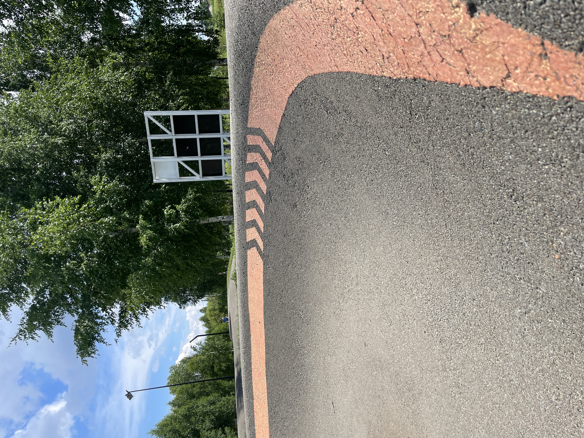 This is a low-angle, outdoor, daylight photograph of a paved area with orange markings. In the foreground, a dark gray paved surface curves from the left to the right of the frame. Along the edge of this paved area, there is a prominent bright orange marking that follows the curve. This orange marking is composed of dashed chevron shapes pointing towards the right.
In the background, the paved surface continues, rising into a gentle hill. Further back, a white structure with black square sections is visible, framed by the leaves of green trees. The trees are lush and dense, with some of their trunks visible. The sky above is blue with scattered white clouds.
A person wearing a blue shirt is visible in the middle ground, standing on the paved area. Other indistinct figures and structures are present in the distance. The overall environment suggests an outdoor recreational space, possibly a park or a track. The image appears to be taken in Laishevo, Russia, given the context.