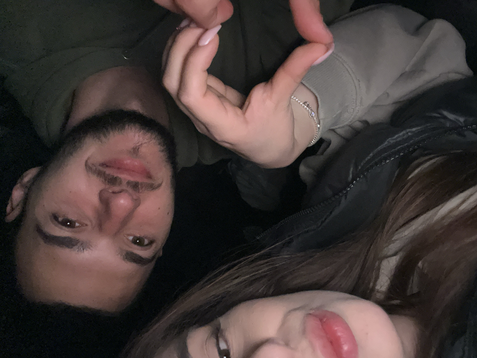 Two people are closely posed together, facing the camera, creating a selfie-style image. One person, likely male, has dark hair, a beard, and brown eyes. The other, likely female, has brown hair and is wearing makeup. Their hands are positioned together to form a heart shape. The male is wearing a green hoodie, while the female is wearing a light-colored hoodie and what appears to be a black puffer jacket. The background is dark and indistinct. The location in Plovdiv, Bulgaria, is not discernible.