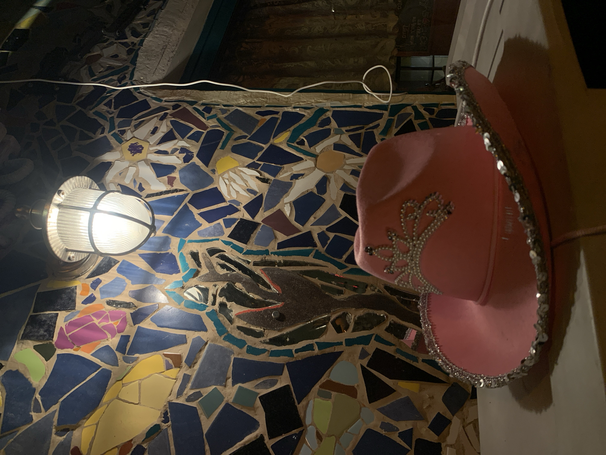 An overhead, close-up shot captures a bright pink, sequined cowboy hat resting on a flat, light-colored surface. The hat is decorated with a tiara-like embellishment on its front and a sequined band around its brim. Behind the hat, a vibrant mosaic wall dominates the scene. The mosaic is composed of irregularly shaped tiles in various shades of blue, yellow, green, and purple, with some darker, almost black, tiles interspersed. A shiny, metallic mosaic piece resembling a flame or a stylized plant is embedded in the center of the wall. A ribbed, glass light fixture hangs from the ceiling above the wall, casting a warm glow on the mosaic. A white electrical cord snakes down the wall to the right. The background suggests an indoor setting with a glimpse of a window with a dark curtain. The overall impression is that of a whimsical and decorative space.