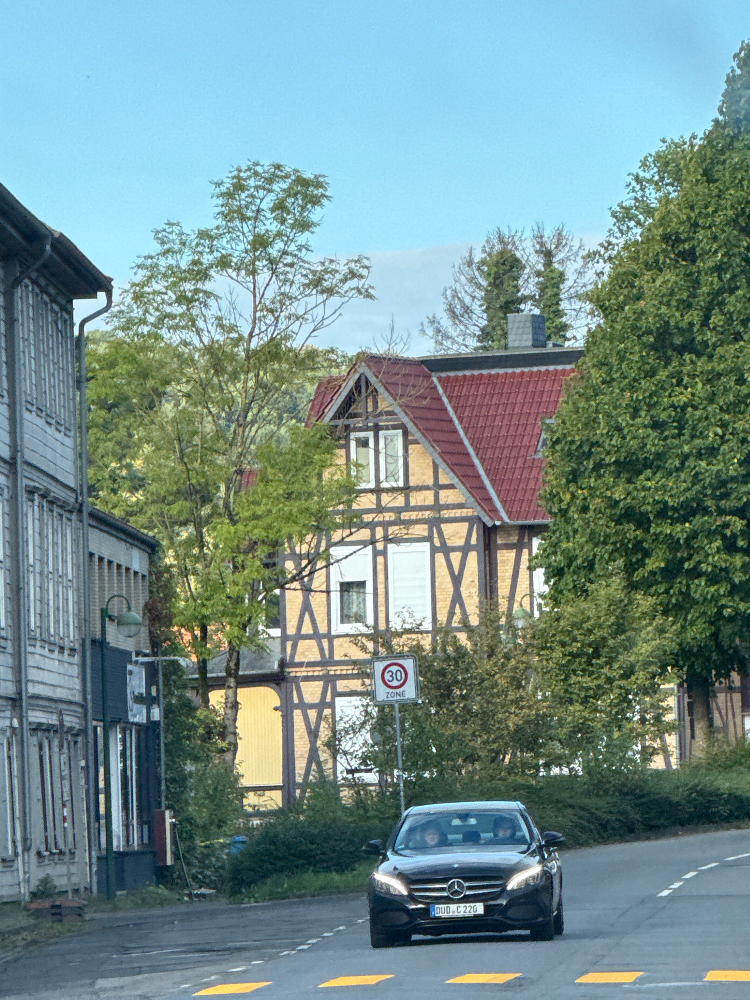 In Osterode am Harz, Germany, a black Mercedes-Benz sedan with license plate DUD C 220 drives on a paved road. Two individuals are visible inside the car. The road is marked with yellow speed bumps and a white dashed line. A red circular sign with "30 ZONE" indicates a speed limit. To the left of the road, a grey building with multiple windows and a dark grey gutter is seen. Further down the street, a building with yellow siding, timber framing, and a red roof stands prominently. Dense green trees and foliage surround the buildings and extend into the background, with a clear blue sky overhead.