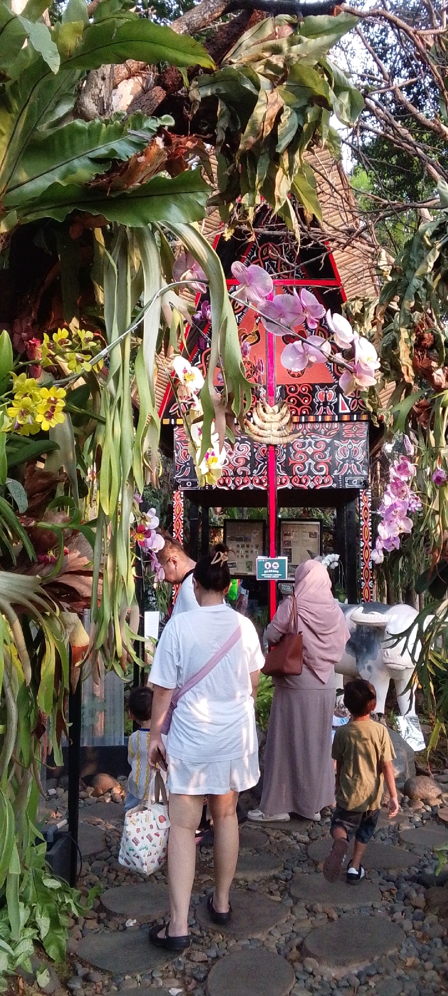 The image depicts an outdoor scene featuring a decorative structure adorned with traditional patterns and surrounded by lush foliage and orchids. In the foreground, several individuals are visible. A woman in a white top and shorts, carrying a patterned bag, walks away from the camera. Beside her, a young boy in a green shirt and patterned shorts also walks away. To the right, a woman wearing a pink hijab and a long dress carries a brown handbag. Another individual, partially visible, stands behind them. A statue of a white bull or water buffalo is positioned to the right of the structure. The ground is covered with stones and stepping stones. The overall setting suggests a cultural or garden exhibit, possibly in an urban park or tourist attraction within Central Jakarta, Indonesia.