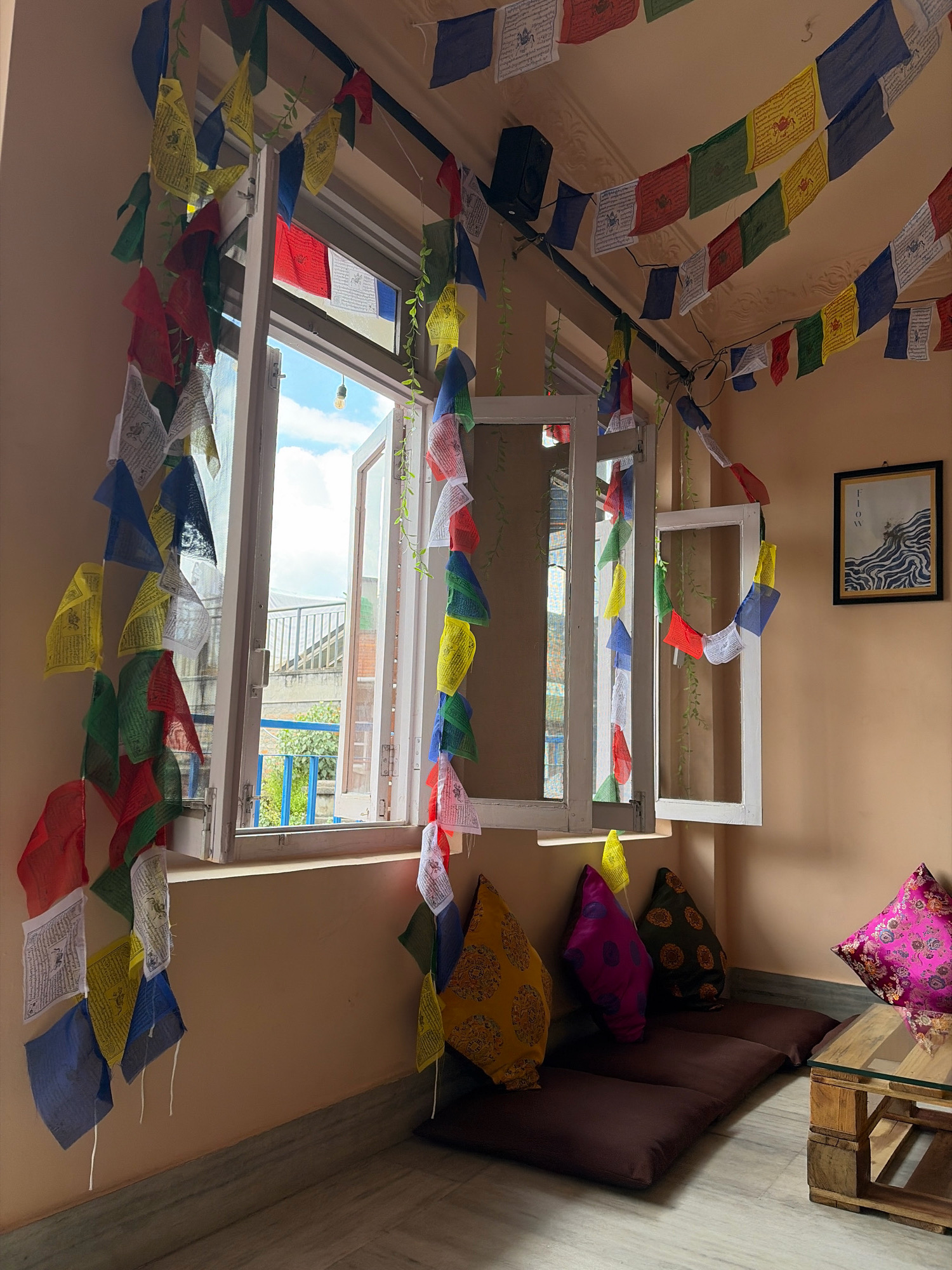 This image depicts an interior scene, likely a room in Lalitpur, Nepal, characterized by vibrant prayer flags. The flags, in various colors including blue, red, green, yellow, and white, are strung across the ceiling and hang down along a window. The window itself is open, revealing a glimpse of an outdoor space with a railing and some greenery. Inside, the walls are painted a warm, light orange hue.
In the foreground, several floor cushions are arranged, topped with decorative pillows. One pillow is a bright yellow with a patterned design, another is purple with pink polka dots, and a third is dark green with an orange circular pattern. To the right, a low, rustic coffee table made of wooden pallets supports a glass top. A framed piece of art, featuring black and white wavy lines against a white background, hangs on the wall above the coffee table. The floor is tiled with a light grey and white pattern. The overall impression is of a cozy, culturally rich space.