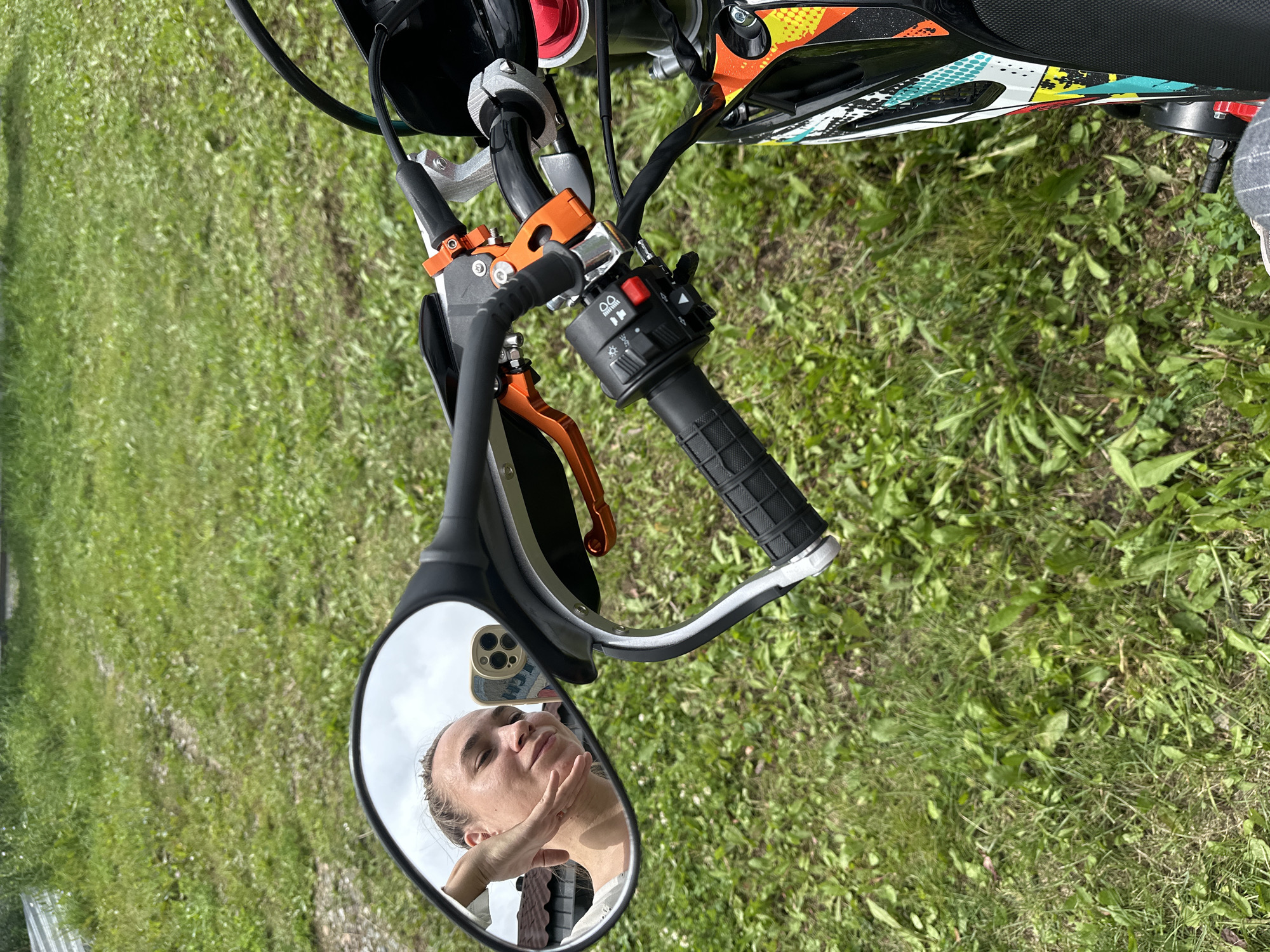 A woman is reflected in the mirror of a motorcycle parked on a grassy area. She is resting her head on her hand and smiling. The motorcycle is black with colorful graphics and orange accents. The background is a blur of green grass and trees. The scene appears to be a rural area, possibly a park or a backyard. The location is not specified as Kharnavy, Russia.