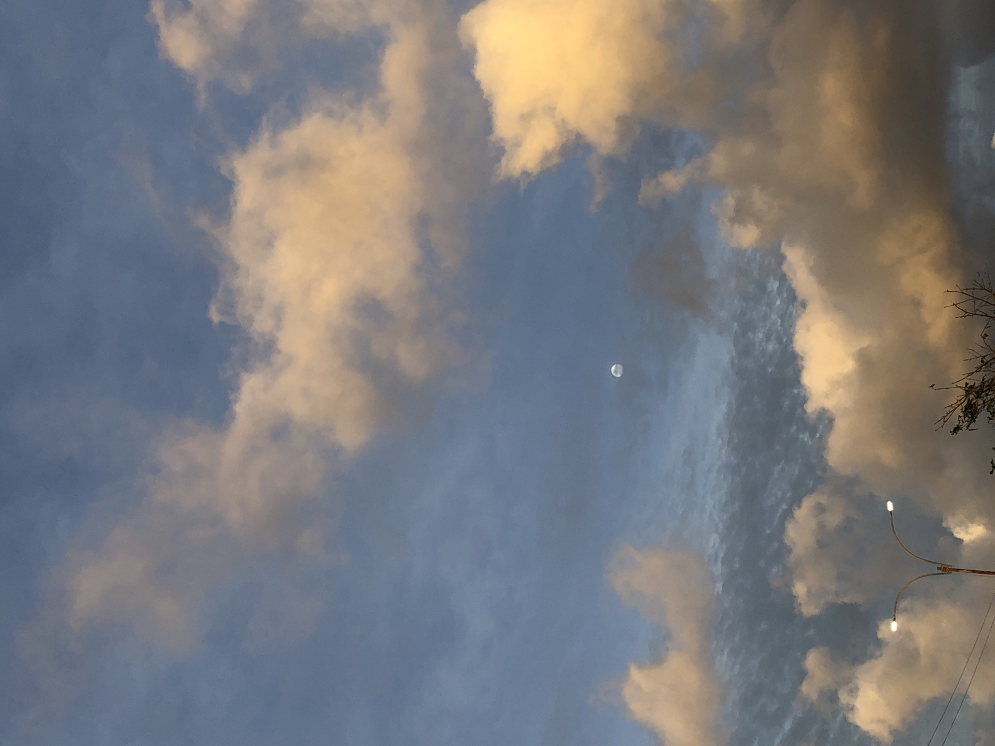 The image depicts a daytime sky with scattered clouds, a faint moon, and the top of a streetlight pole with two illuminated lights. The clouds are illuminated by the sun, displaying shades of yellow and white. The sky is a soft blue. The top of the streetlight pole is visible on the right side of the image. The location is Juan Griego, Venezuela, as indicated in the prompt. There are no people present in the image.