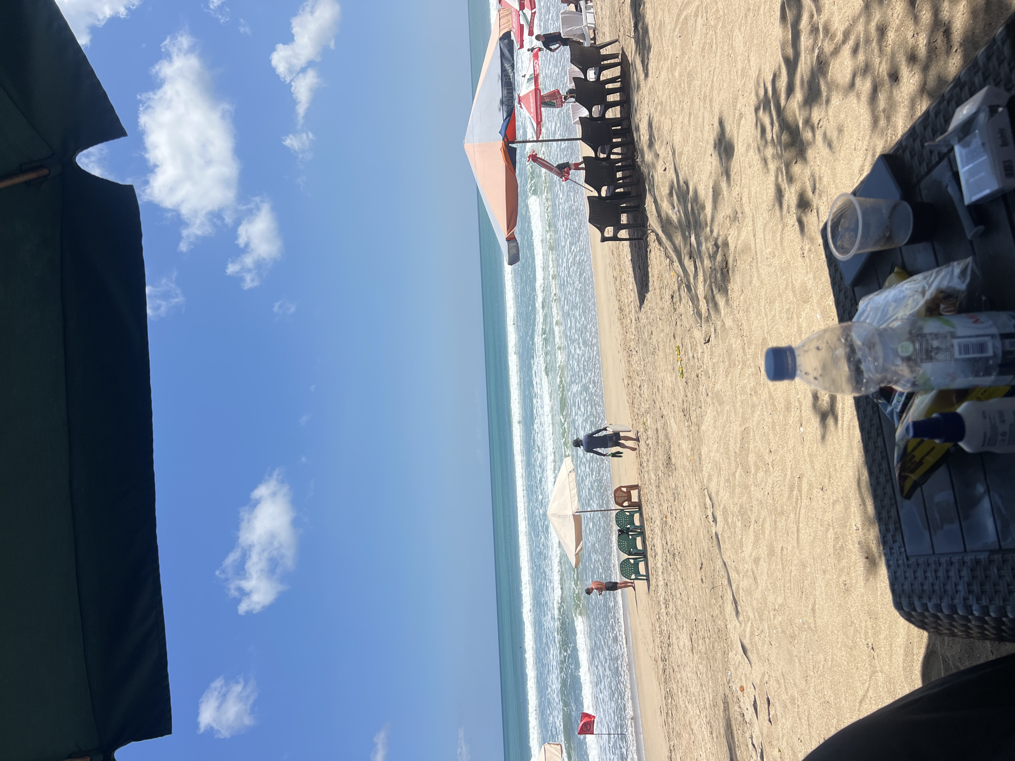 The image depicts a sunny day at Kuta Beach, Bali, Indonesia. In the foreground, a dark woven table is partially visible, holding a plastic bottle of water, a plastic cup with a dark liquid, a smartphone, and a pack of cigarettes. The table casts a shadow onto the sandy beach. In the mid-ground, several beach chairs are arranged under large umbrellas. One umbrella is orange and white, while another is beige. A red flag with a circular symbol is planted in the sand near the water. Two men are visible on the beach; one is standing near the beige umbrella, looking at his phone, and another is walking towards the ocean carrying a surfboard. The ocean has moderate waves, and the sky is a clear blue with scattered white clouds. The general atmosphere suggests a relaxed beach setting.