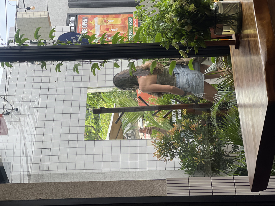 The image is a reflection in a window. The reflection shows a person with long, dark hair, wearing a green top and denim shorts. They are holding a phone, seemingly taking a picture. Behind them is a white tiled wall with a rectangular mirror reflecting greenery. Lush green plants are present both inside and outside, obscuring parts of the scene. The image appears to be taken from inside a building, with a wooden table and plants in view. There are advertisements visible on the building across the street.