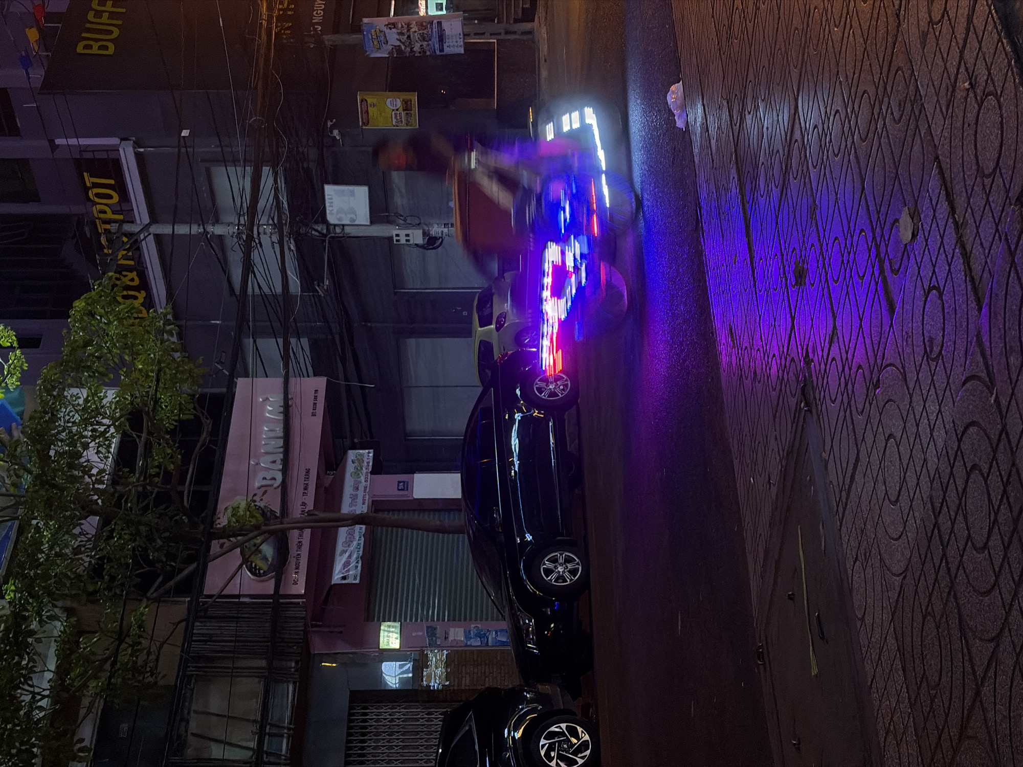 A long exposure photograph captures a nighttime street scene in Nha Trang, Vietnam. A three-wheeled vehicle with illuminated sides, emitting streaks of blue and purple light due to motion blur, is the central subject. The vehicle is traveling on a paved street, reflecting the colorful lights and the surrounding environment. To the left of the vehicle, two black cars are parked. The building in the background features signage for "BÀNH MÌ" (Vietnamese for "bread"), indicating a food establishment. Other signs and wires are visible, typical of a city street. The ground is a patterned sidewalk with some debris. The overall lighting is dim, with the vehicle's lights and building signage providing the primary illumination.