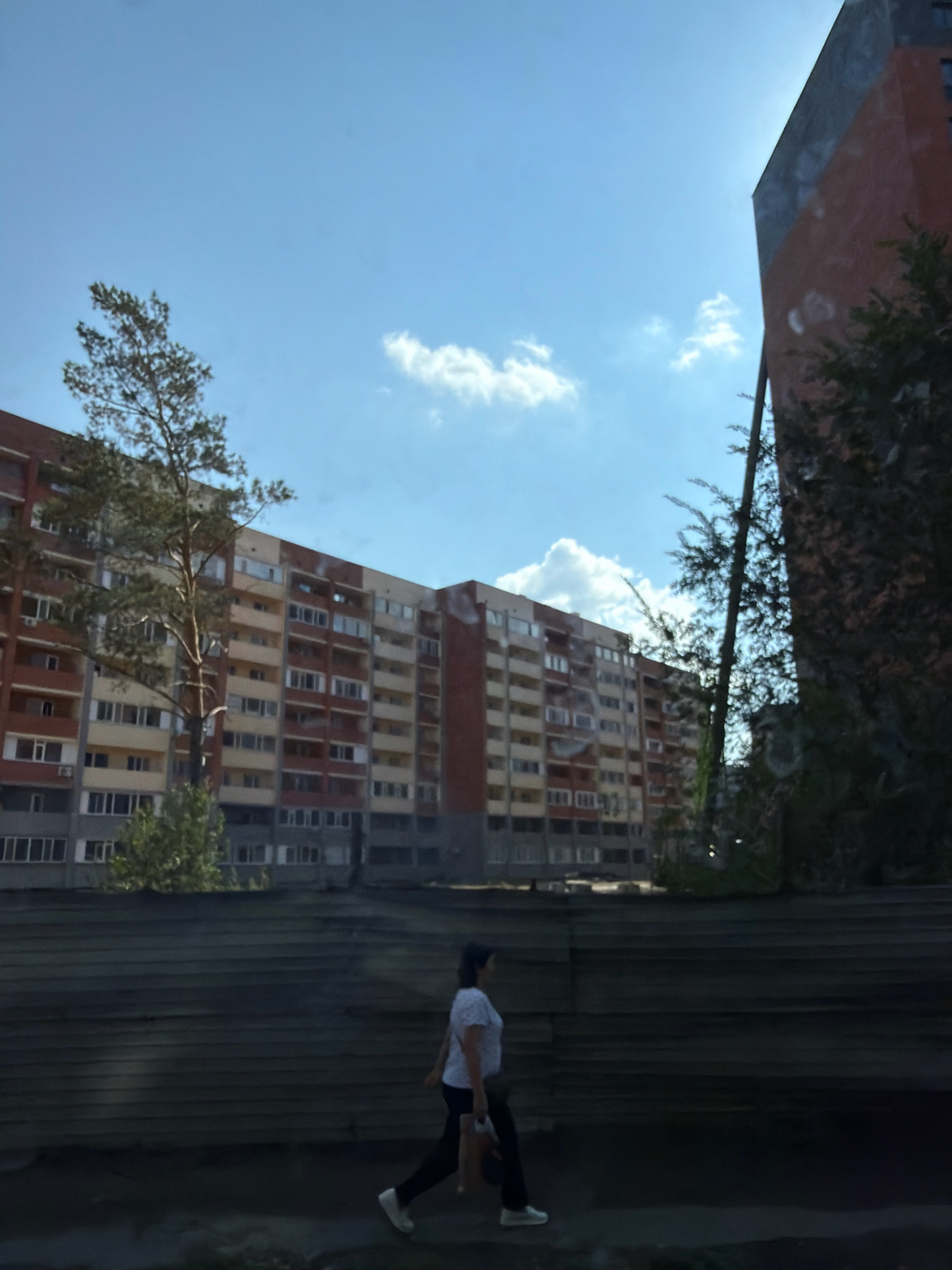 An urban scene in Pavlodar, Kazakhstan, is captured under a bright, partly cloudy sky. In the foreground, a woman walks from left to right, wearing a white short-sleeved shirt and black pants. She carries a light-colored bag in her right hand. She is walking on a dark, textured surface that appears to be a pathway or sidewalk. Behind her, a low, dark, horizontal structure with a wood-grain texture stretches across the frame, acting as a visual barrier.
In the middle ground, a large, multi-story apartment building dominates the scene. It is a long, flat-roofed structure with numerous balconies and windows, exhibiting a color palette of light yellow, beige, and red brick. A tall evergreen tree stands prominently in front of the building, its branches reaching towards the sky. To the right of the apartment building, a taller, modern building with a red brick facade and a dark upper section is visible. Lush green trees and foliage surround the base of this building and extend into the background. The overall environment suggests a residential area within the city.