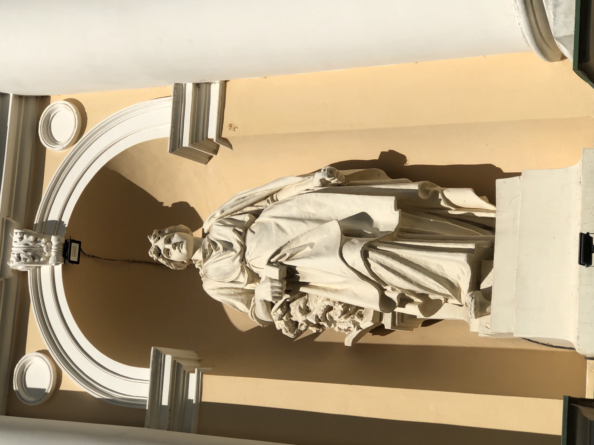 This image captures a statue of a robed female figure standing within an arched alcove of a building. The statue is made of light-colored stone and is positioned slightly off-center to the right. The figure is draped in flowing garments and holds an object in her left hand. To her left, partially obscured, is a sculpted bust of a bearded male head. The architectural elements of the alcove are white, contrasting with the peach-colored wall behind the statue. Sunlight casts sharp shadows on the wall and the statue, indicating a bright, sunny day. The statue appears to be part of the architectural facade of a building, likely a public structure or a historical monument. This statue is located in Lviv, Ukraine, specifically at the National Opera House of Lviv.