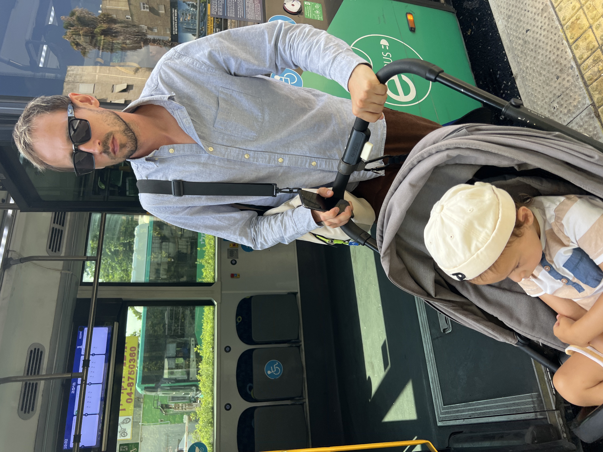 A man wearing sunglasses and a light blue button-down shirt stands next to a stroller containing a baby. He is holding the stroller handle and appears to be boarding or disembarking from a bus. The bus is green and white, with the word "BUS" visible on its side, along with a white "e" in a circle. There are seats inside the bus, some marked with a blue wheelchair symbol. The background shows buildings and palm trees, suggesting an urban environment. The scene takes place on a sunny day at a bus stop in Kiryat Bialik, Israel, as indicated by the presence of a digital display inside the bus showing Hebrew text and a time of 10:02.