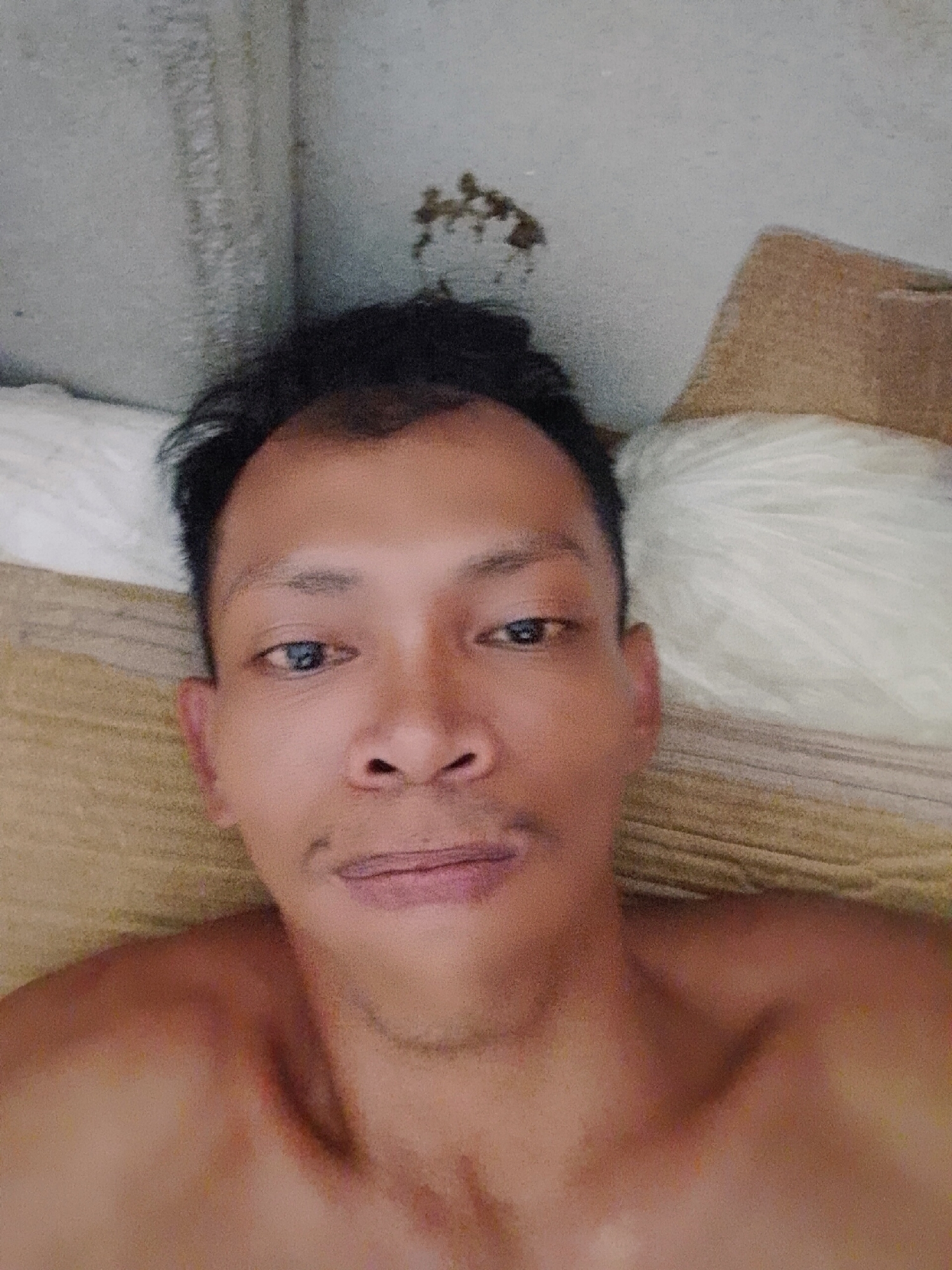 The image is a selfie of a man lying down. He has dark hair and appears to be shirtless. He is positioned on what looks like a bed or couch with various pillows. The background includes a wall with some discoloration. The location is specified as Bukit Ketok, Indonesia.