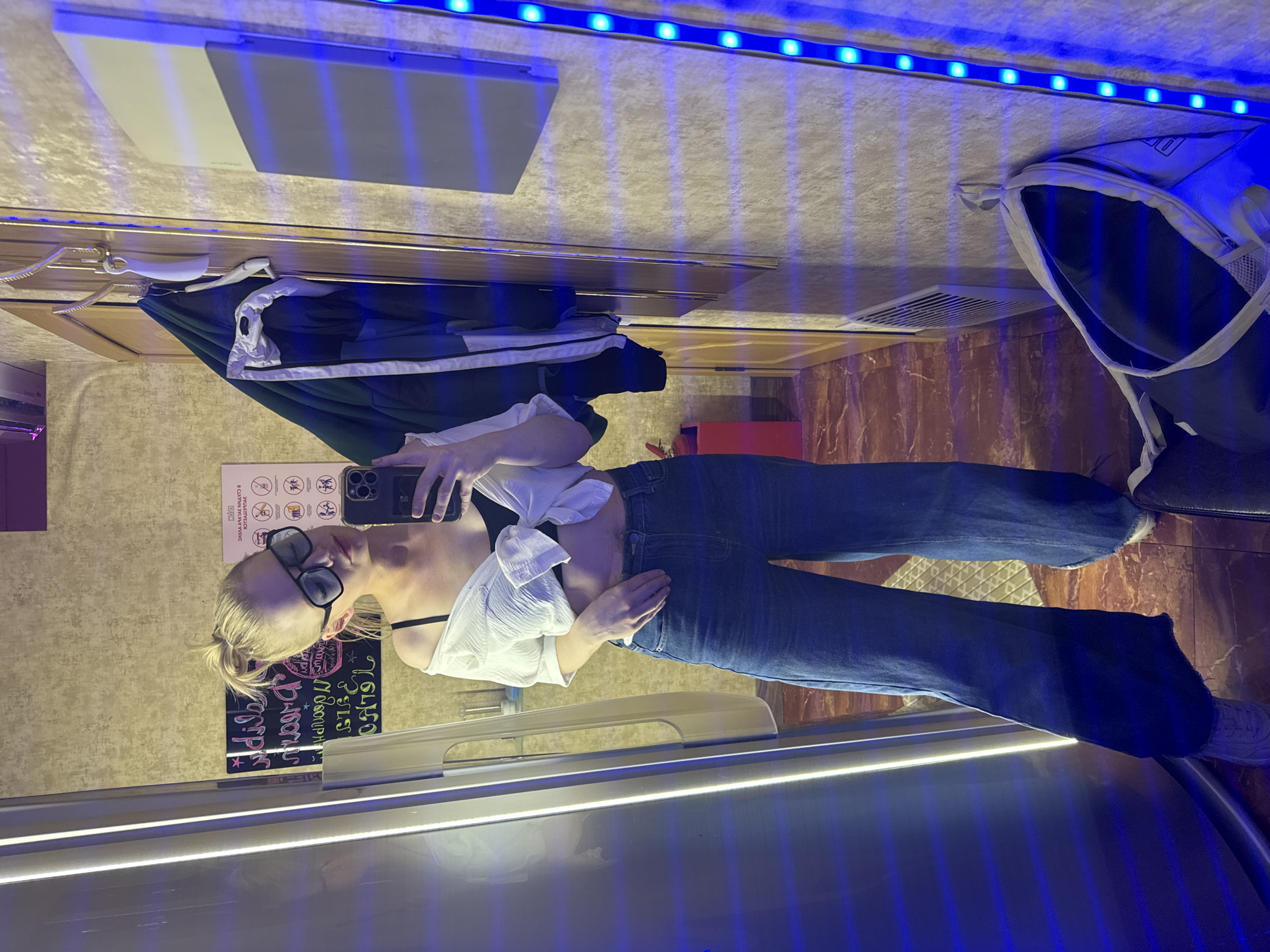 A woman takes a mirror selfie indoors. The location has wood paneling, clothes hanging from a rod, and a poster with symbols on the wall. The woman is wearing jeans, a black bra, and a white shirt that's tied at the waist. She is also wearing sunglasses and has a blonde ponytail. Blue LED lights are visible at the top and bottom of the frame. The photograph does not contain enough information to determine that it was taken in St. Petersburg, Russia.
