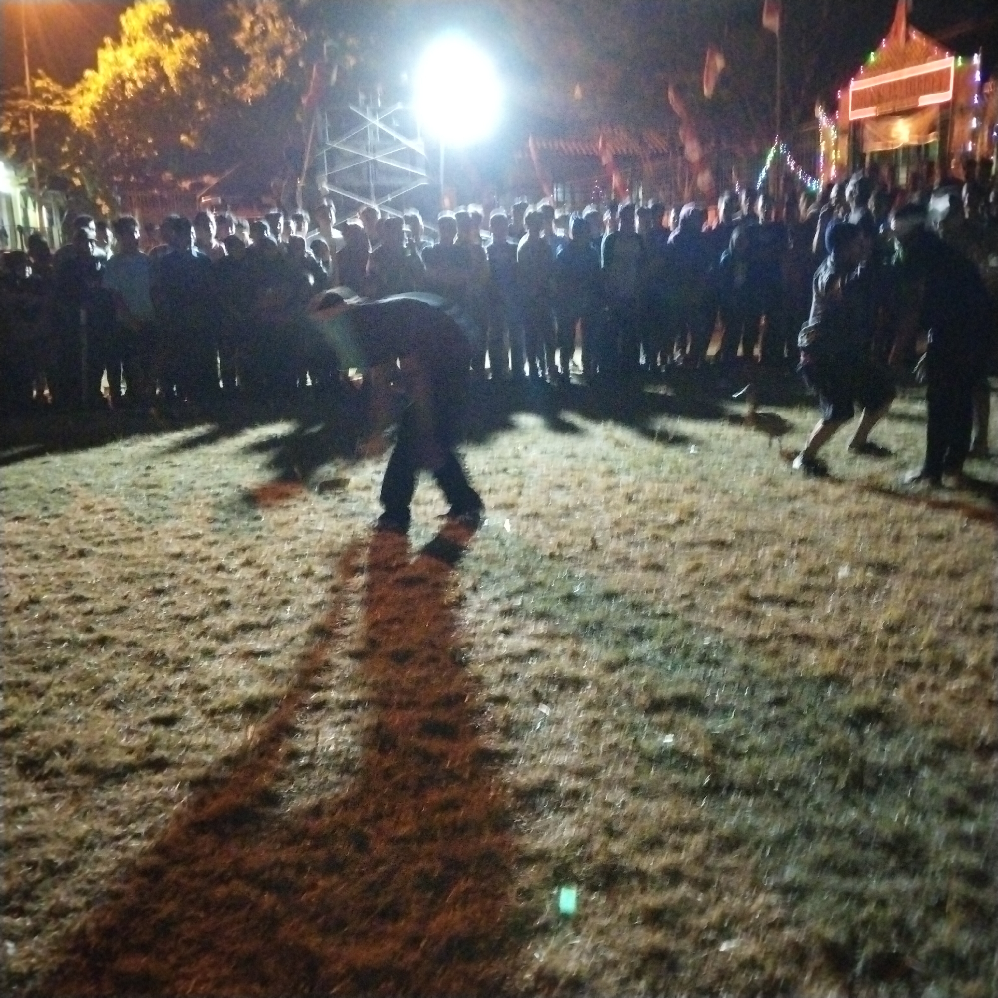 The image depicts a nighttime scene in Pamulihan, Indonesia, likely an outdoor gathering or event. A crowd of people is visible, mostly silhouetted against bright lights. The ground appears to be covered in dry grass or earth, with distinct shadows cast by the figures and structures. In the foreground, a person is bent over, their shadow stretching prominently across the ground, suggesting they might be engaged in an activity. In the background, a large, bright light source is prominent, possibly a spotlight or a large lantern, illuminating a structure that appears to be a stage or a decorated building adorned with lights. The overall atmosphere suggests a festive or communal event taking place after dark.