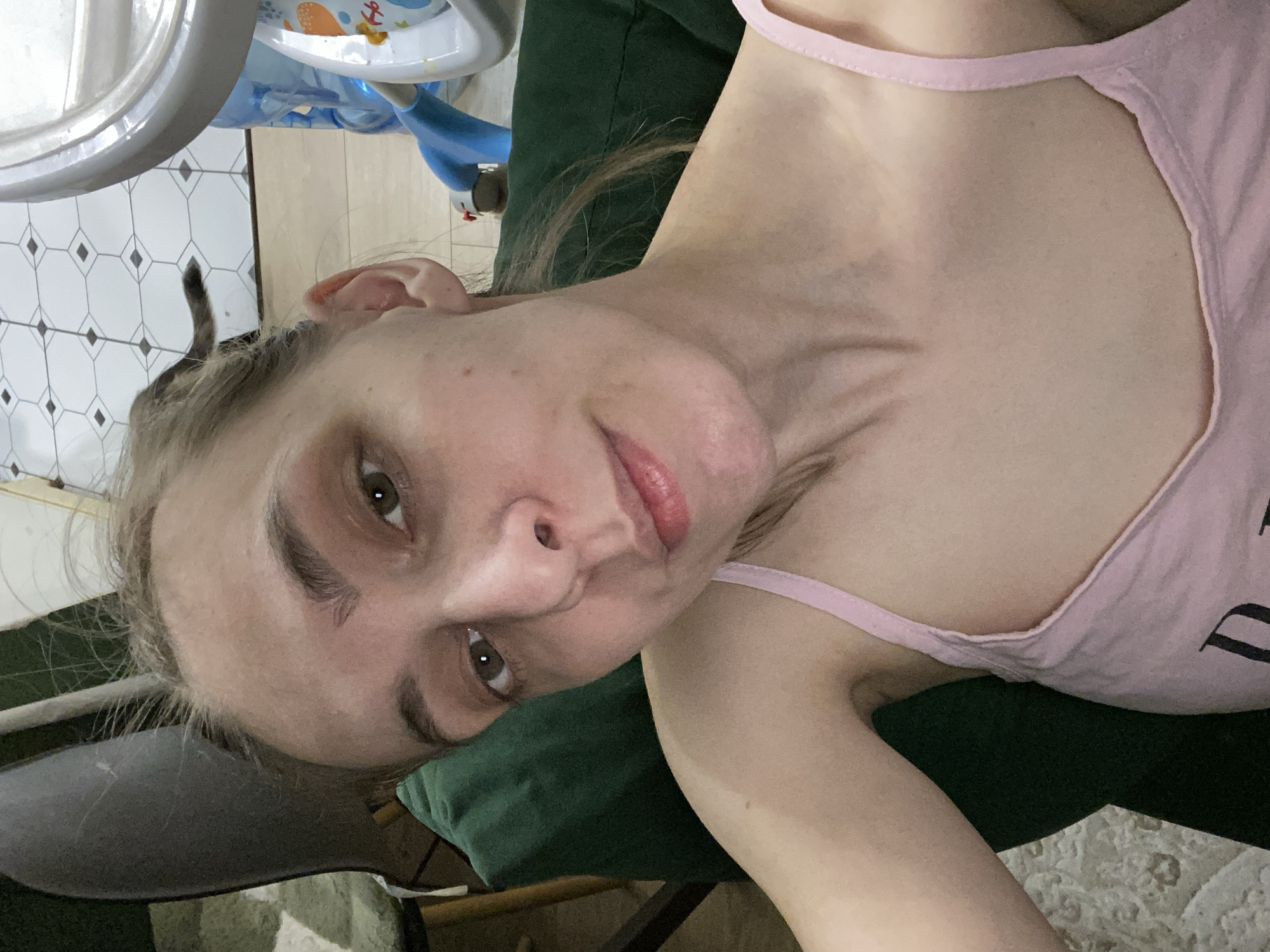 A young adult female, light-skinned with light brown hair pulled back, appears in a close-up selfie in an indoor domestic setting in Chaikovsky, Russia. She wears a light pink camisole and looks directly at the camera with a neutral expression. Her right arm is partially visible, suggesting she is holding the camera. The background to the left features a dark green upholstered surface and a wooden furniture leg, above a light-colored patterned rug. To the right, a white tiled floor with a dark geometric pattern is visible, along with parts of a blue and white plastic baby high chair. A dark grey cat's tail is visible extending from behind the woman's head towards the tiled floor.