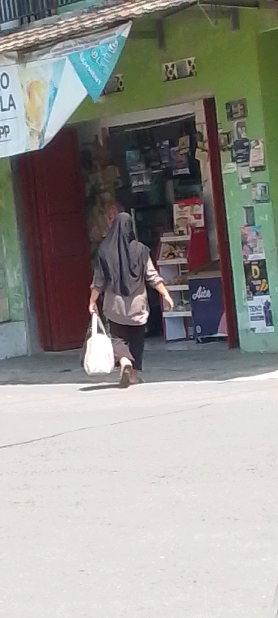 A woman wearing a dark hijab and a light-colored tunic with dark pants walks away from a storefront in Cilaku, Indonesia. She carries a light-colored tote bag in her left hand. The storefront has a red door and a green exterior wall adorned with posters and a decorative window grill. A triangular banner with blue and white graphics is attached to the building's facade above the entrance. The ground in front of the store is a paved road or sidewalk.