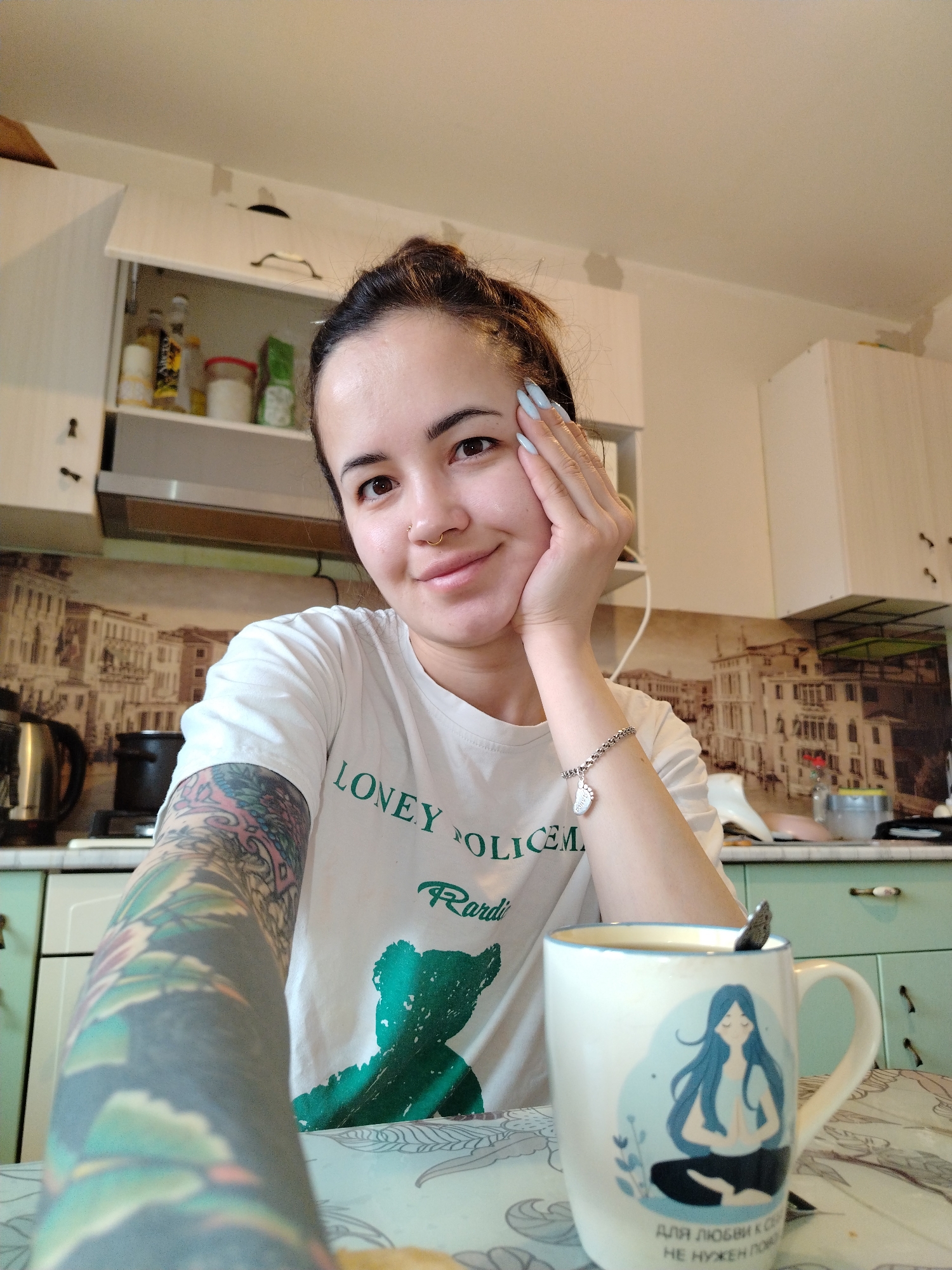 A young adult woman is captured in a selfie, sitting at a table in a home kitchen, likely in Noviy, Russia. She smiles gently, resting her chin on her right hand, which has light blue nail polish. Her dark hair is pulled back, and she wears a small nose ring. Her left arm is extensively covered in colorful, intricate tattoos, forming a full sleeve. She wears a white short-sleeved t-shirt featuring partially visible green text "LONEY POLICEM Rardi" and a green silhouette of a bear. On her right wrist is a silver bracelet with a small charm, possibly shaped like a footprint.
In the foreground, a light-colored mug with a blue illustration of a meditating woman sits on a patterned tablecloth. Visible Cyrillic text on the mug reads: "ДЛЯ ЛЮБВИ К СЕБЕ НЕ НУЖЕН ПОВОД," which translates to "FOR SELF-LOVE, NO REASON IS NEEDED." A spoon is placed inside the mug, suggesting she is enjoying a drink.
The background reveals a domestic kitchen setting with white upper cabinets and light green lower cabinets. One upper cabinet is slightly ajar, showing various bottles and containers. A stainless steel kettle, a pot, and an oven hood are visible on the counter. The backsplash features a wallpaper depicting detailed buildings, reminiscent of a European cityscape. The overall impression is of a relaxed, everyday moment in a cozy home environment during daytime.