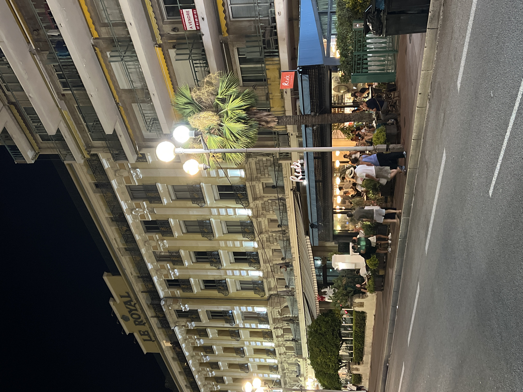 The image depicts a nighttime street scene in Nice, France, likely on the Promenade des Anglais, given the presence of palm trees, a large hotel, and a seaside ambiance. In the foreground, a street is visible with white lane markings. The middle ground shows a sidewalk bustling with activity. A hotel, identified by a sign reading "LE ROYAL," dominates the background with its illuminated facade, showcasing classical architectural elements like columns and balconies with ornate railings. To the right of the hotel, another building with multiple balconies, some with yellow awnings, is visible. A sign advertising "A VENDRE" (For Sale) is attached to this building.
On the sidewalk, several people are seen walking and dining at outdoor tables. A prominent palm tree stands near the hotel's entrance. Restaurants with awnings and outdoor seating occupy the lower level of the hotel and adjacent buildings. The overall atmosphere suggests a lively evening in a popular tourist area.
