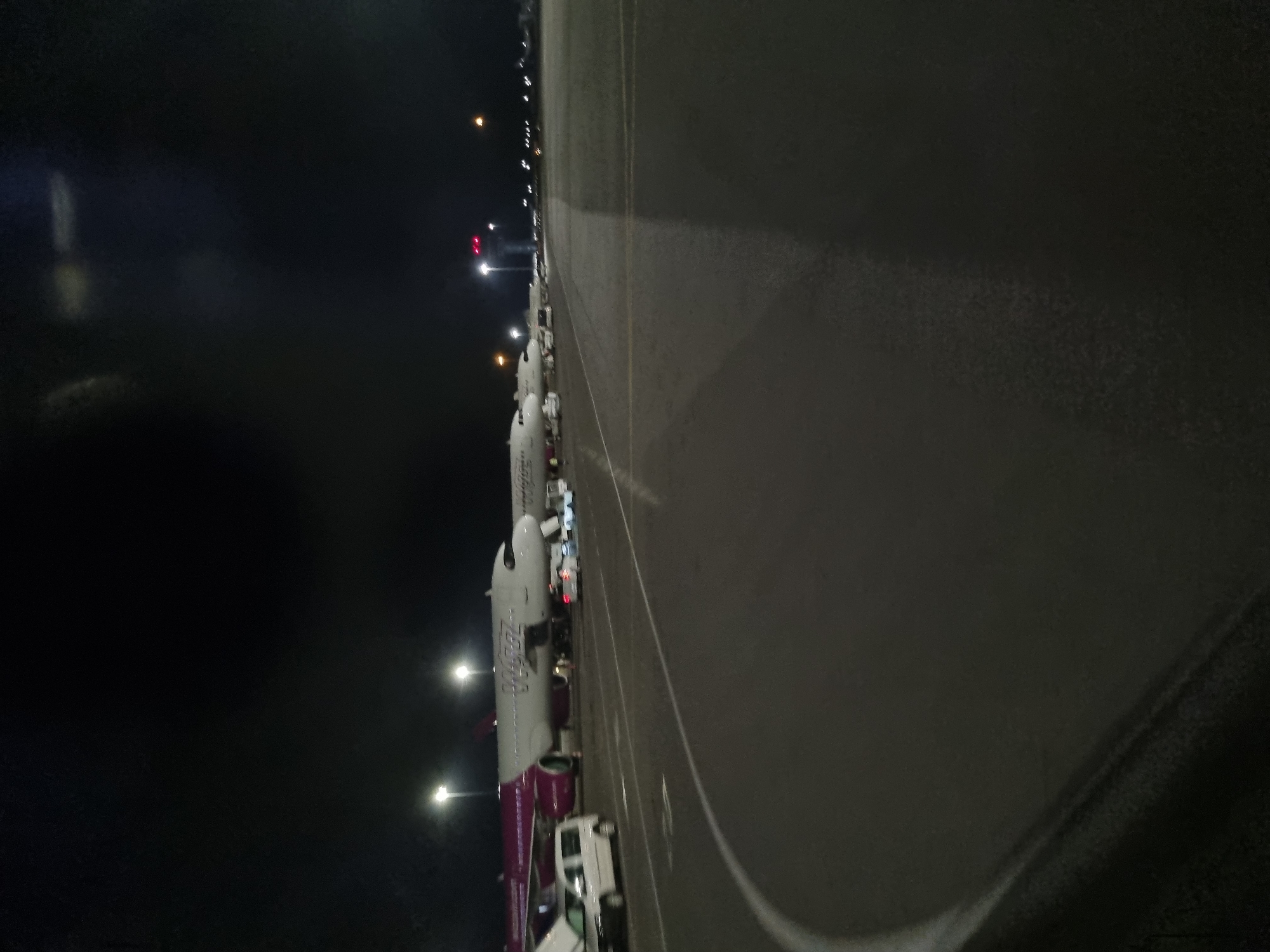 At night, the image depicts a row of parked airplanes along an airport tarmac, likely at Sofia Airport. The planes are mostly white with colored markings, including purple and red. Service vehicles, including vans, are parked near the planes. The tarmac is dark gray, and the environment is lit by sparse streetlights and vehicle headlights. The sky is dark, and visibility is somewhat limited. The exact position in Sofia is not discernable.