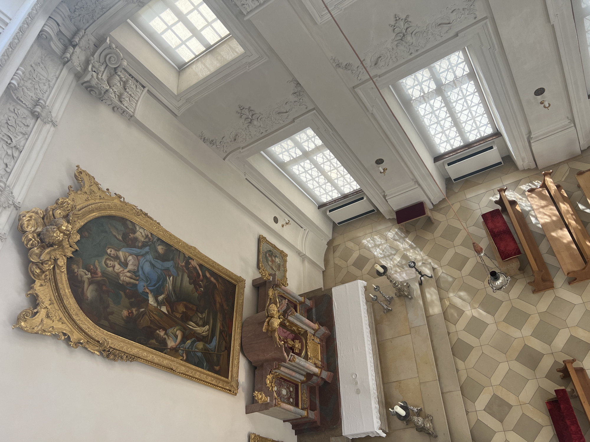 The photograph provides an overhead view of the interior of the St. Georgen church in Oberschleißheim, Germany, looking towards the altar area and surrounding architecture. Dominating the upper portion of the image is a large, ornate gold-framed painting depicting a religious scene, likely the Madonna and Child with Saints, suspended from the ceiling. Below this, a richly decorated altar with gold and marble accents is visible, covered partially by a white cloth.

The floor is tiled in a geometric pattern of beige and gray squares. Several wooden pews, some with red velvet cushions, are arranged in the foreground and to the right. On the left side of the altar area, a set of stone steps leads up. Various decorative elements, including candlesticks, are placed on the floor and steps.

The walls are white and feature intricate stucco work, particularly around the windows and cornices. Two large windows with ornate grilles are visible on the right side, allowing natural light to filter into the space. The overall impression is one of opulent Baroque or Rococo church architecture. No people are visible in the frame.