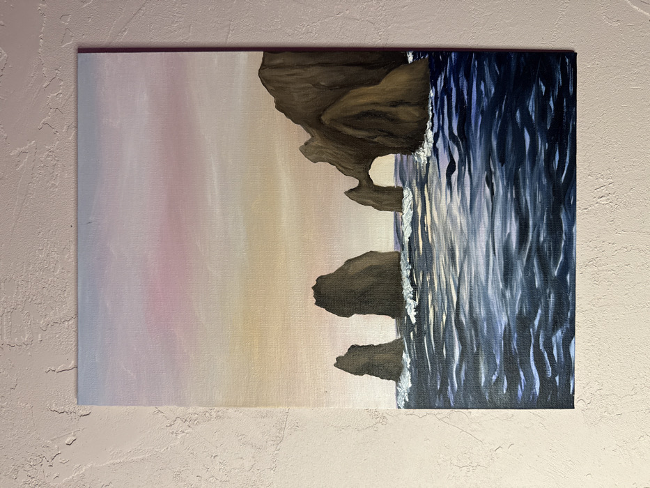 This image presents a vertical painting on canvas, physically situated in Rostov-on-Don, Russia. The artwork depicts a dynamic seascape at either sunrise or sunset. The sky above transitions smoothly from a pale blue at the zenith, through soft pink mid-tones, to a warm golden-yellow hue at the horizon line. Below the horizon, a deep blue ocean is rendered with visible waves and white foam breaking against several dark brown rock formations. On the left side of the painting, three distinct sea stacks rise prominently from the water. On the right, a larger, more complex rock mass features a well-defined natural archway. The painting itself is mounted directly onto a light-colored, textured wall. No individuals, man-made objects, or specific interactions are visible within the depicted natural environment.