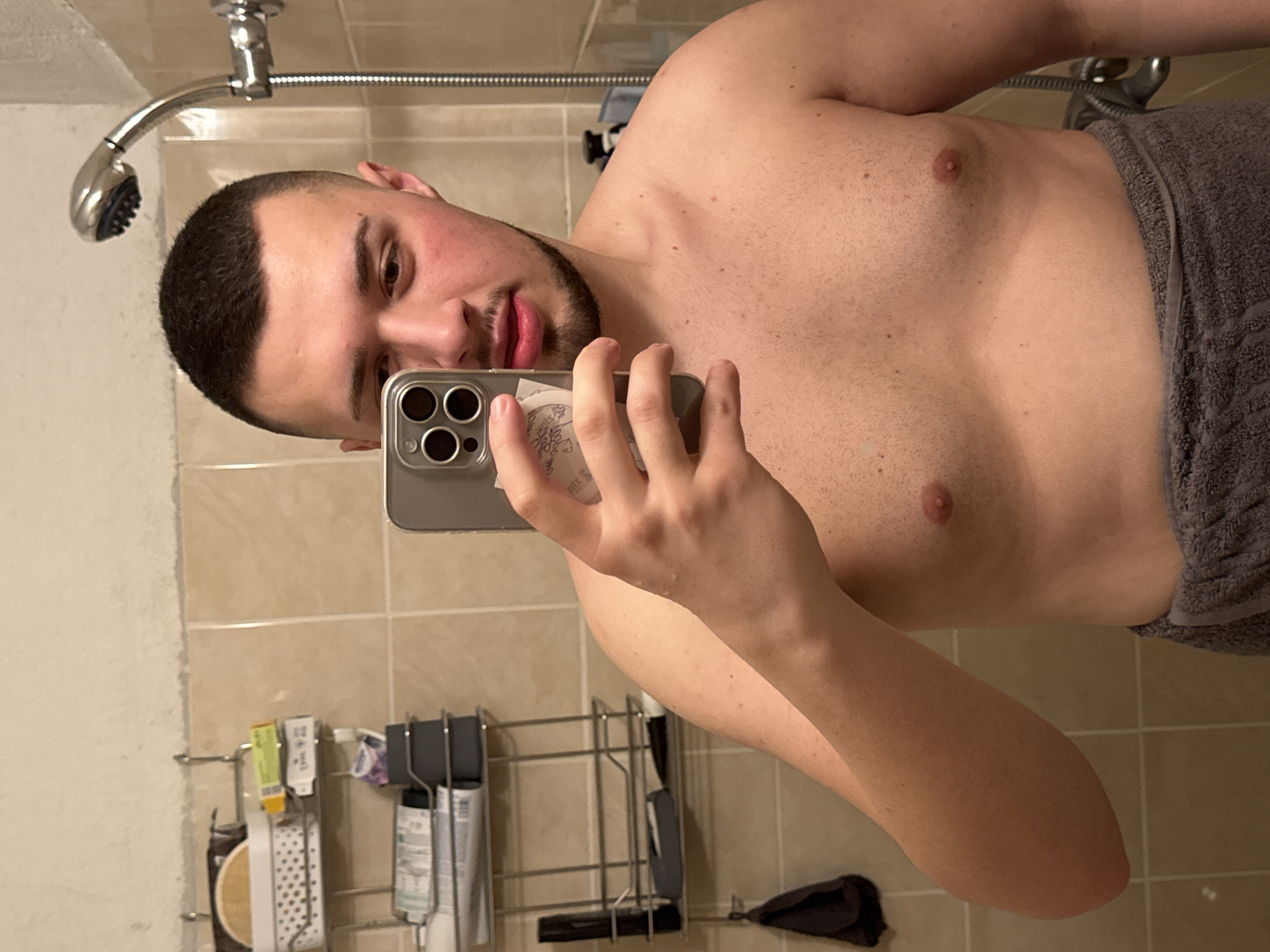 The image shows a shirtless man in a bathroom taking a selfie with his phone. He has short dark hair, a beard, and a fair complexion. He is partially wrapped in a grey towel. The background features beige bathroom tiles, a showerhead, and a rack holding toiletries. There is no evidence to suggest this photo was taken in Sofia, Bulgaria.