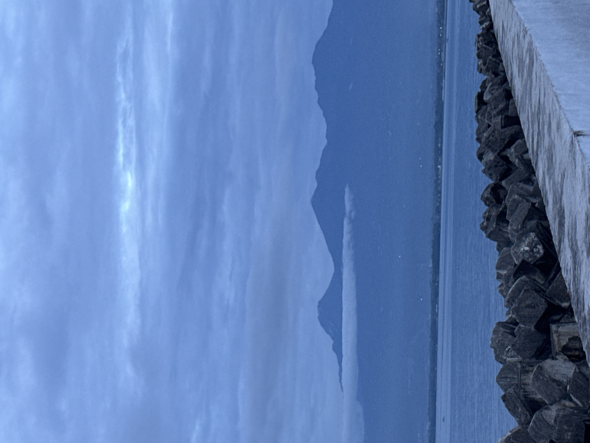 The image depicts a landscape with a body of water, possibly a lake or sea, stretching horizontally across the frame. On the right side, there is a man-made structure, likely a concrete wall or breakwater, constructed with dark-colored, irregularly shaped rocks. In the distance, a mountain range is visible, its peaks shrouded in a blue-gray haze. The sky is overcast with a layer of clouds, giving the scene a muted, monochromatic feel. The city of Davao City, Philippines cannot be ascertained from this image.