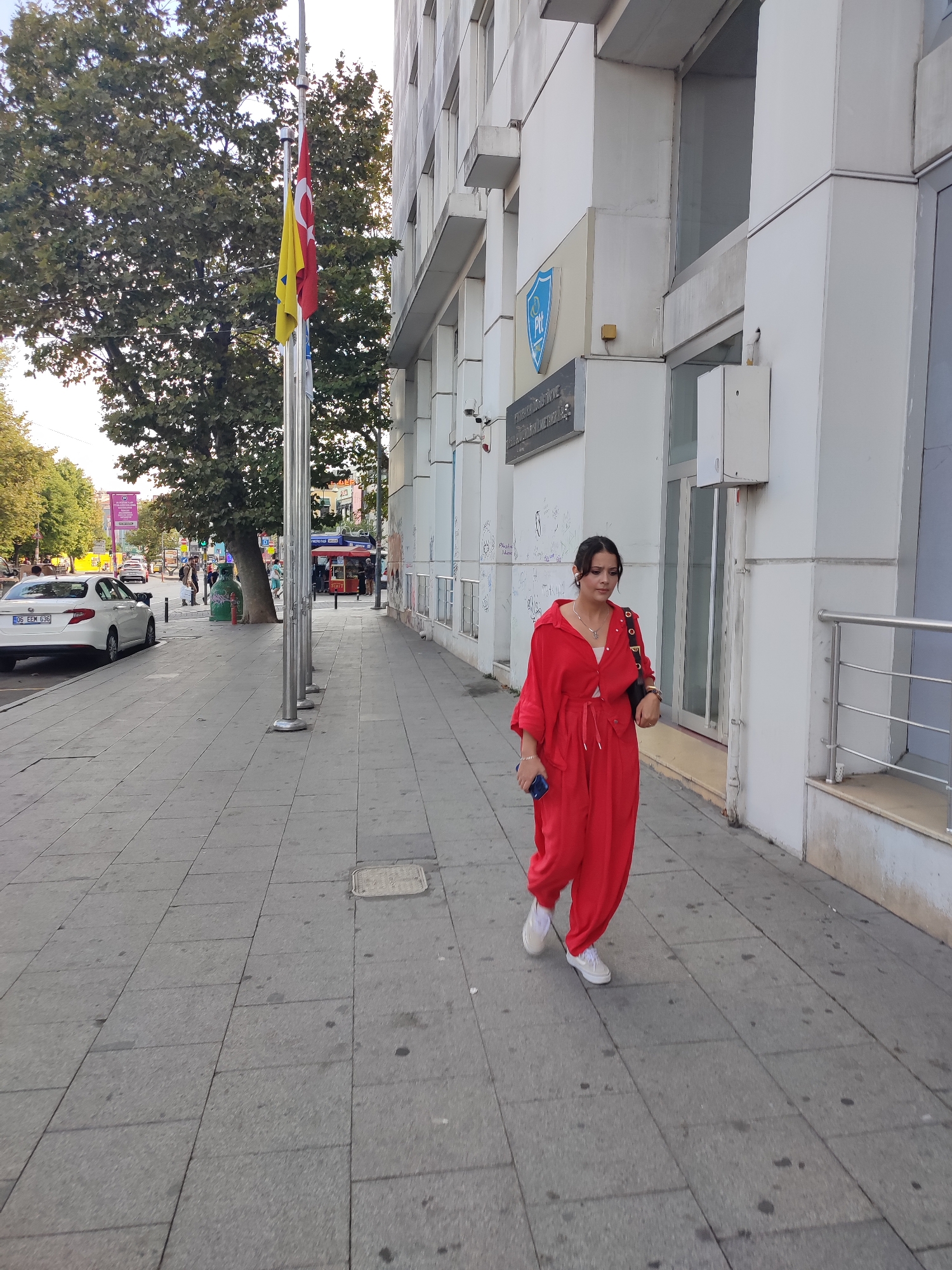 A young woman wearing a red jumpsuit and white sneakers walks down a sidewalk in front of a modern white building. She is holding a blue smartphone in her left hand and has a black purse slung over her right shoulder. The building has large glass windows and several signs, including one with a blue shield logo and text that appears to be in Turkish. To the left of the building, a street is visible with cars and trees lining the sidewalk. Two flags, one red with a white star and crescent and another yellow, are flying from a flagpole in front of the building. The sidewalk is made of grey concrete tiles. The scene appears to be in Kadıköy, Turkey, a district in Istanbul known for its commercial and residential areas.
