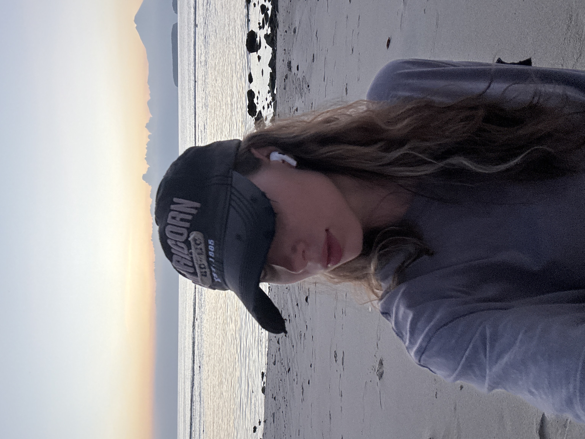 A young woman wearing a black baseball cap and a gray t-shirt stands on a sandy beach at sunset. She has light brown wavy hair and is wearing white earbuds. The background shows the ocean with calm waves and a faint outline of mountains in the distance, typical of the Kertasari, Indonesia coastline. The sky is a soft gradient of orange, pink, and blue.