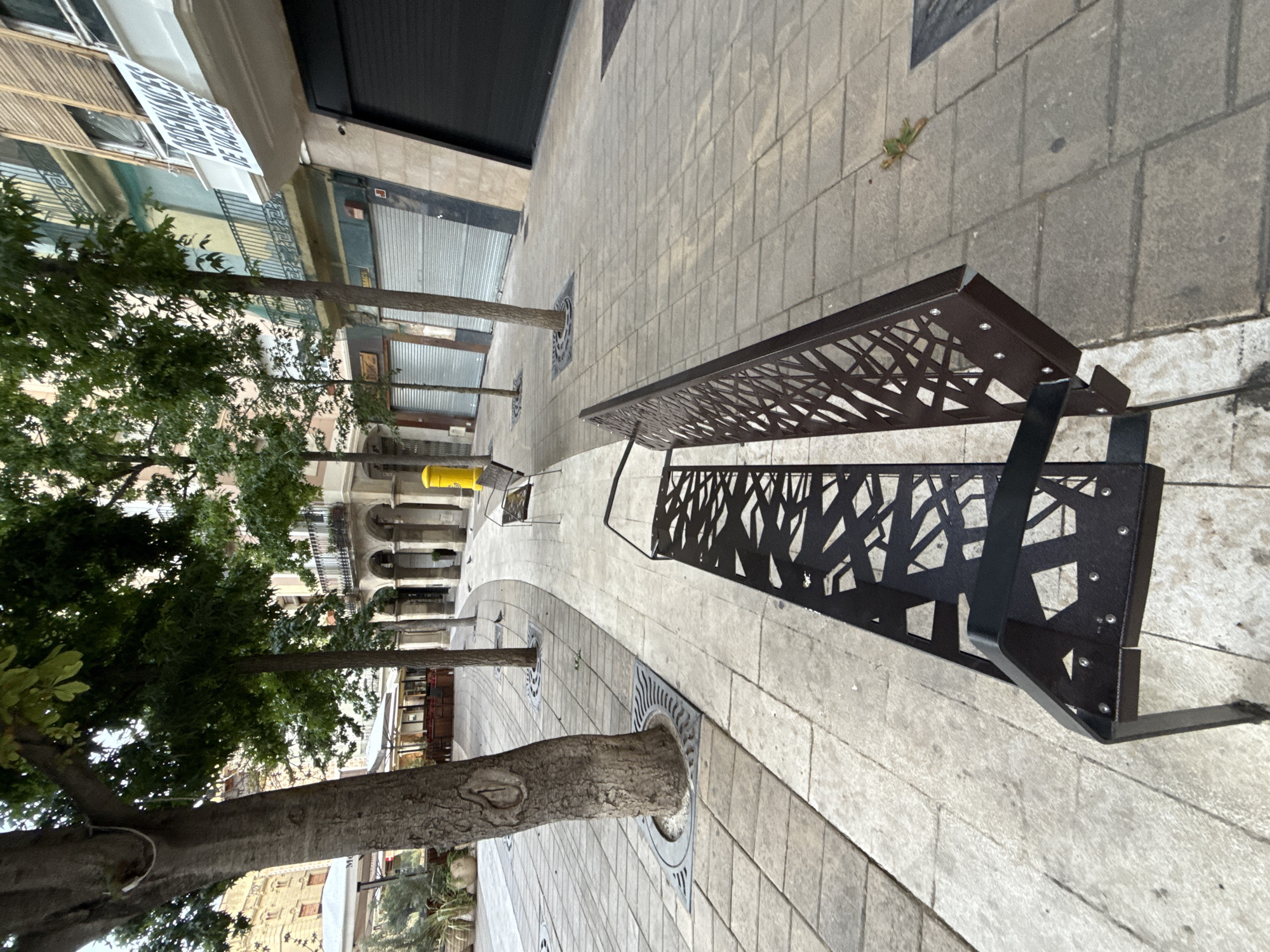 This is a wide-angle, ground-level shot of a pedestrian walkway in Igualada, Spain, with a modern black metal bench in the foreground. The bench has a geometric, cutout design on its backrest and seat. The walkway is paved with light-colored rectangular tiles.
To the left, large trees with rough bark are planted in circular grates with a wave pattern. Beyond the trees, there are white umbrellas shading outdoor seating areas, possibly for cafes or restaurants, with wooden chairs and tables visible. Further in the background, the facade of a building with arches and balconies can be seen.
To the right, the walkway continues, and another black metal bench is visible in the mid-ground, similar in design to the one in the foreground. A bright yellow cylindrical object, possibly a public bin or bollard, stands near this bench. The buildings lining the right side of the walkway have a colorful facade with balconies and shuttered windows. A white banner with text, including "ORDENANCES DE VACANCES," is hanging from a balcony. The ground level of these buildings appears to be commercial spaces with closed metallic roll-up doors.
The lighting suggests it might be late afternoon or early morning, with shadows cast on the ground from the trees and benches. A few pigeons are scattered on the walkway. The overall scene depicts a clean and well-maintained urban public space.