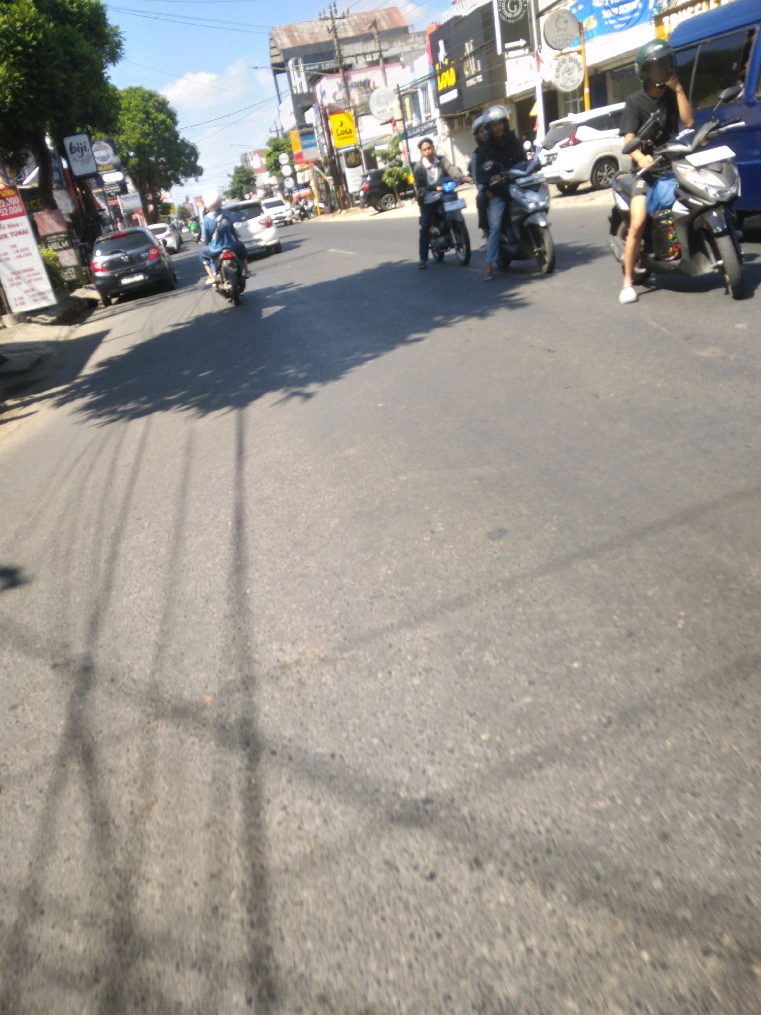 This is a medium-shot, eye-level, outdoor scene on a sunny day, depicting traffic on a road in Sumampir, Indonesia. The foreground is dominated by asphalt, with shadows of trees and utility poles cast across it, indicating a bright, clear sky. In the middle ground, several motorcycles and their riders are visible, moving along the road.
On the right, a male rider wearing a black t-shirt, blue shorts, and a green helmet is on a black and white scooter, looking to his left and talking on a mobile phone. To his left and slightly behind, another motorcycle is occupied by a male rider in a black t-shirt and helmet, and a female passenger wearing a black top and blue jeans. Further to the left, a third motorcycle is ridden by a man in a light-colored jacket and helmet, with a female passenger in a black headscarf and blue jeans.
In the background, white and black cars are parked or driving on the road. Buildings line the right side of the street, featuring signage with Indonesian text, suggesting commercial establishments. A blue van is parked on the far right. The overall scene suggests a typical day with moderate traffic in a built-up area of Sumampir.
