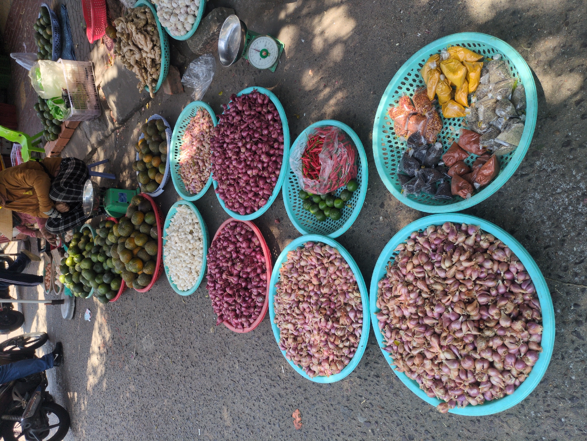 The image depicts a street-side market scene in what appears to be South Quy Nhon Ward, Vietnam. A vendor, dressed in a brown top and checkered pants, is seated next to a variety of produce displayed in blue and red plastic baskets. The produce includes mangoes, garlic, red onions, chilis, limes, ginger, and various spices packaged in small plastic bags. A scale is visible near the produce. A motorcycle and the feet of people walking by can be seen on the edge of the frame, suggesting a busy street. The scene is bathed in bright sunlight.
