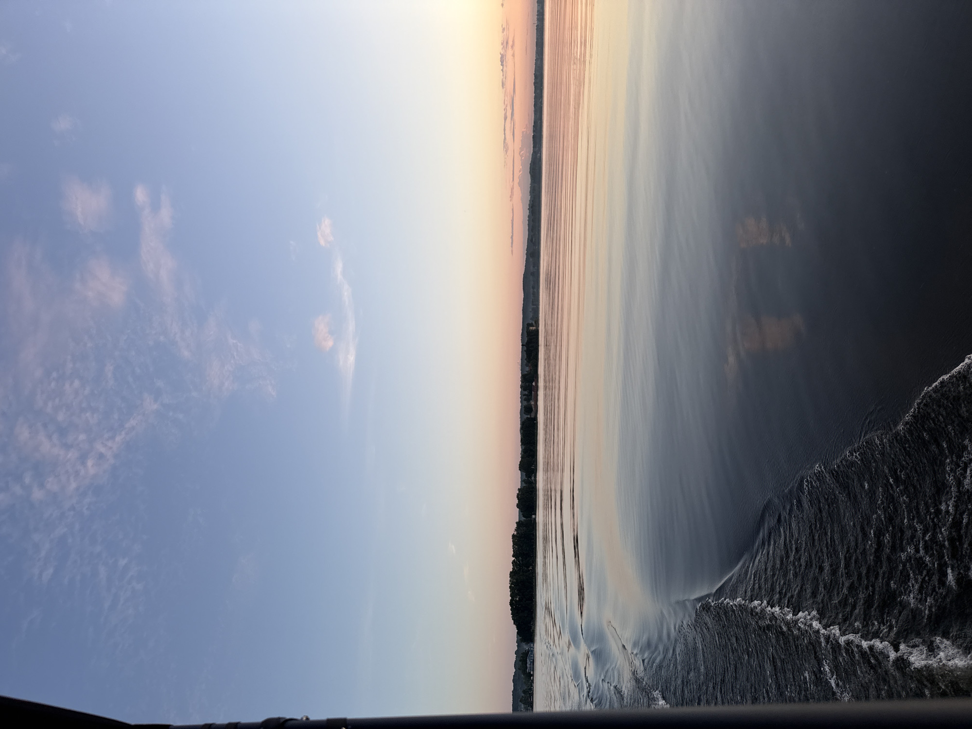The scene, described as being in the city of Votkinsk, Russia, captures a broad body of calm water at either sunrise or sunset. A dark, cylindrical boat rail occupies the left edge of the frame, extending vertically from top to bottom. Adjacent to this rail, a distinct wake of white-capped waves moves across the dark blue-grey water, indicating motion from a vessel. The main body of water is smooth and highly reflective, mirroring the soft gradient of colors in the sky above, which transitions from a light blue at the zenith to pale pinks and oranges near the horizon. Scattered, wispy clouds are visible across the sky, with some reflecting the warm hues of the low sun. Along the distant horizon, a dark, low-lying landmass densely populated by trees is visible, with some indistinct buildings or structures appearing on the far left of the shoreline. Gentle hills or elevated terrain are faintly visible further to the right horizon. No individuals or specific actions are discernible within the frame beyond the implied movement of the boat from which the photograph was taken.